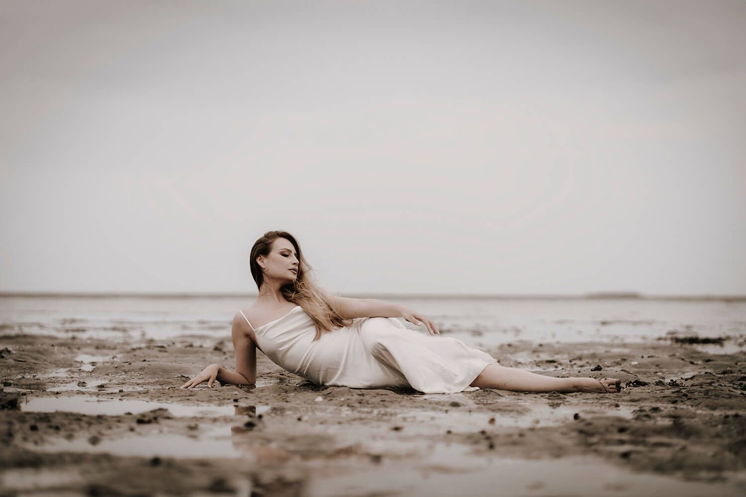 Kvinde i hvid kjole liggende ved vandkanten – æstetisk modelfotografi i Nordjylland.