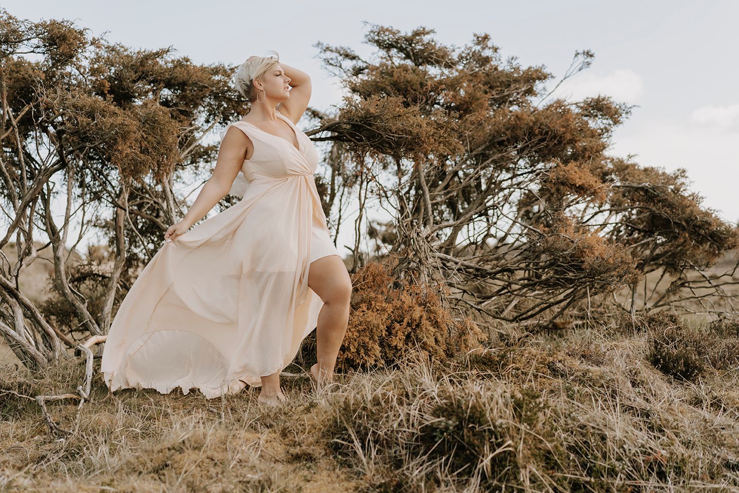 Kvinde med kort blondt hår og lyserød kjole poserer med hånden ved halsen – fotograf Aalborg portræt.