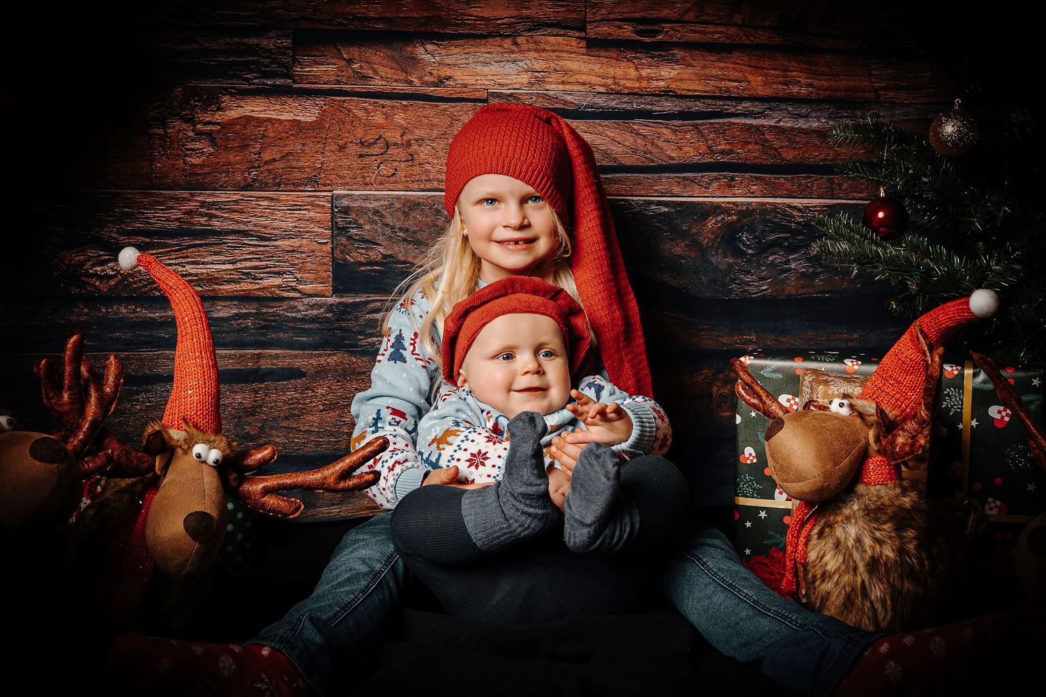 To søskende i nissehuer fotograferet sammen med juletræ, gaver og julepynt.