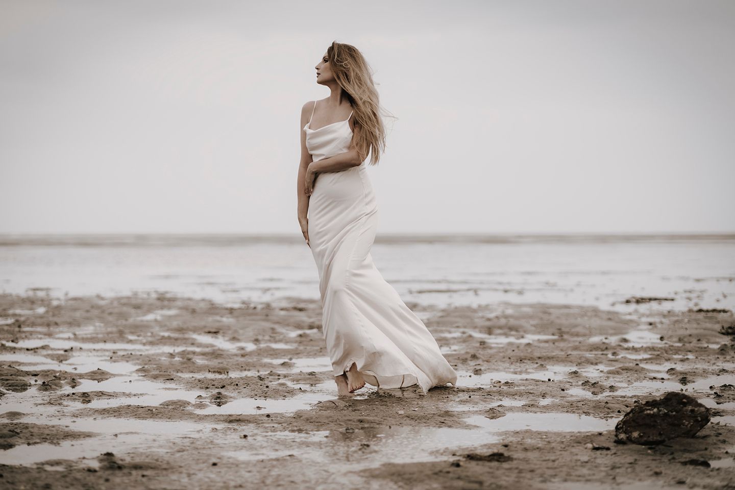 Modelfotograf Aalborg - Kunstnerisk billede af en kvinde i hvid kjole, som går ud over et lavvandsområde ved havet.