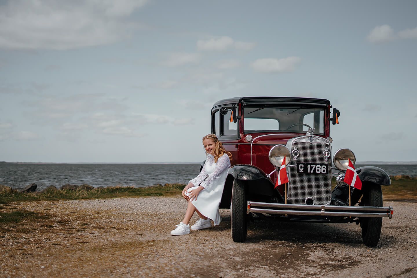 Konfirmationsbillede af pige i kjole fotograferet udendørs ved veteranbil i Nordjylland.