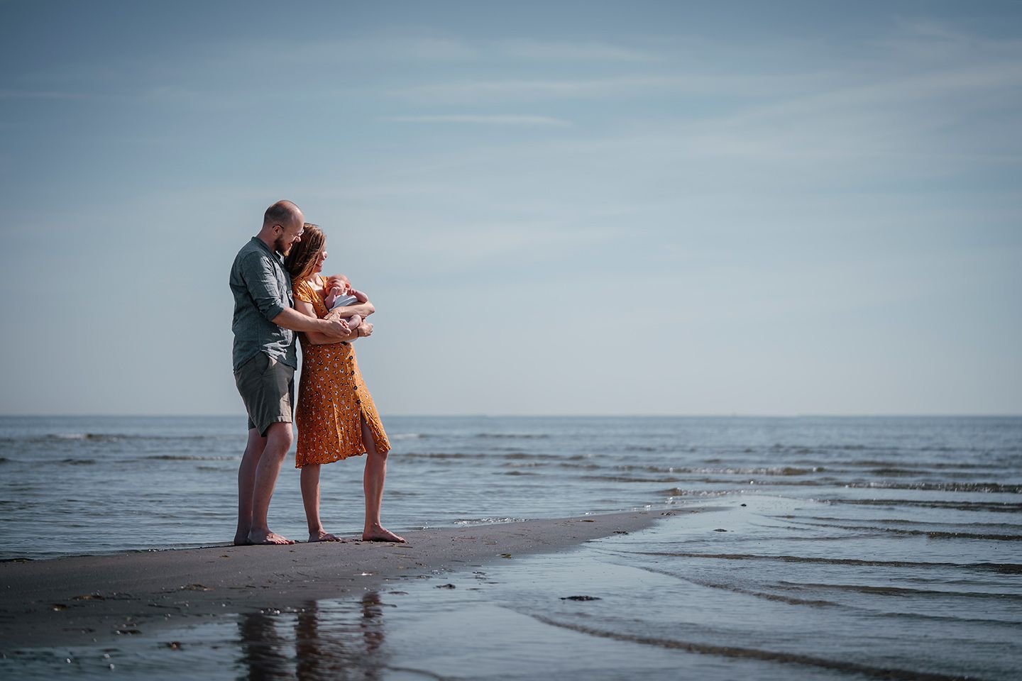 Familiefotograf Aalborg – Et forældrepar står på kanten til havet. Smilende, med deres nyfødte i armene, taget af fotograf i Aalborg, JOEhan