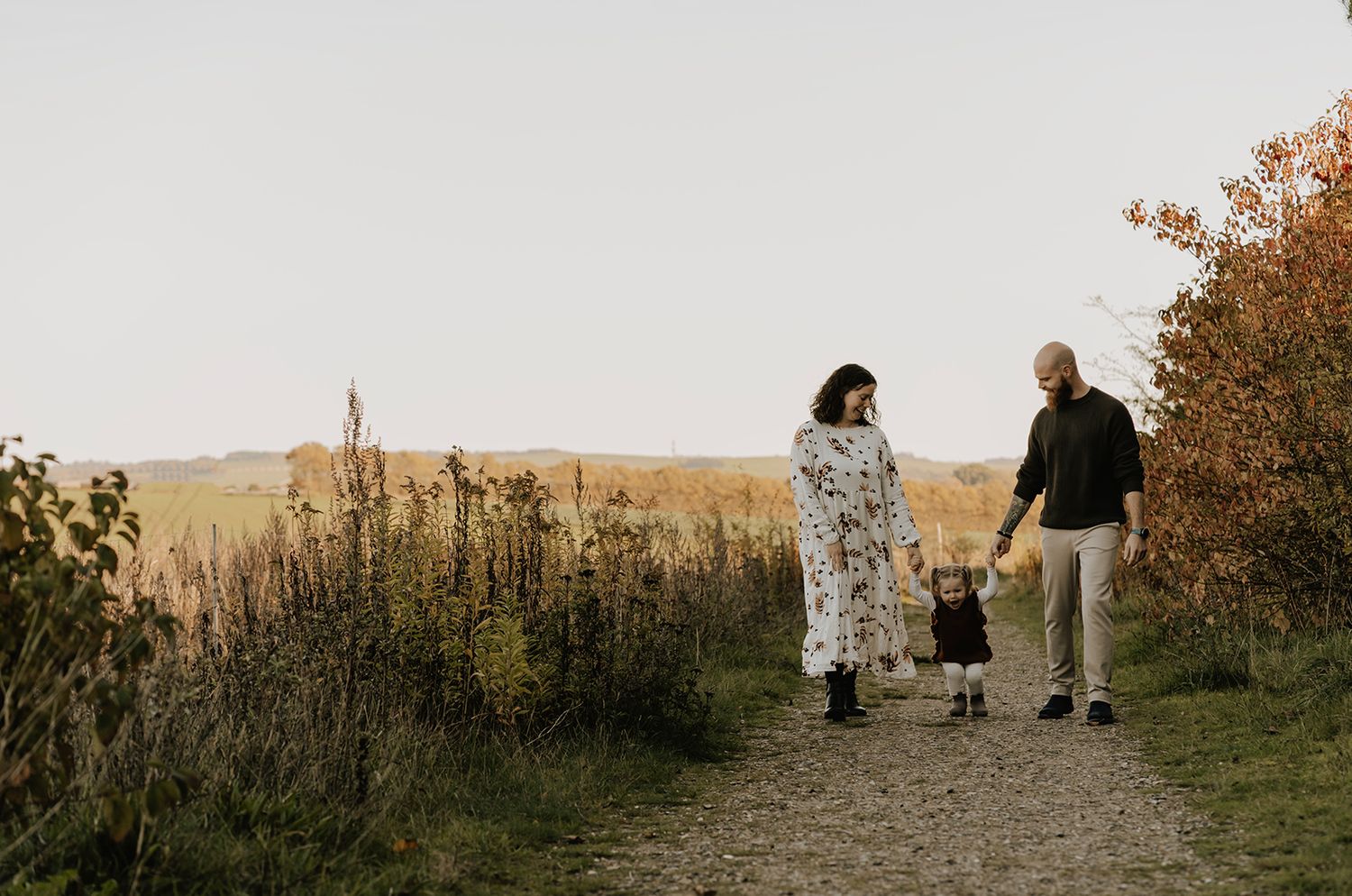 Naturlig familiefotografering i Aalborg og Nordjylland – mor og far går hånd i hånd med deres lille datter på en natursti i efterårets varme