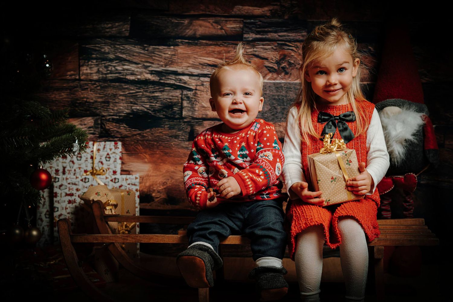 Baby og større søskende fotograferet med juletræ, julegaver og rustik træbaggrund.