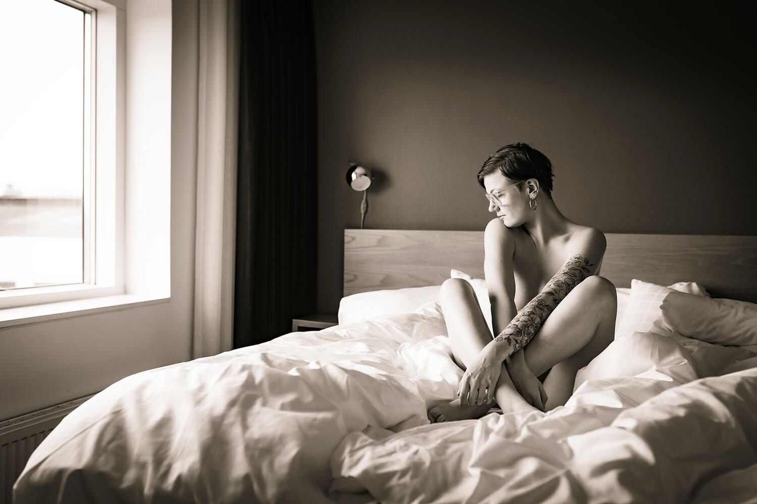 Kvinde med tatoveret arm sidder i sengen og ser ud af vinduet – naturligt boudoirportræt af boudoirfotograf i Aalborg.