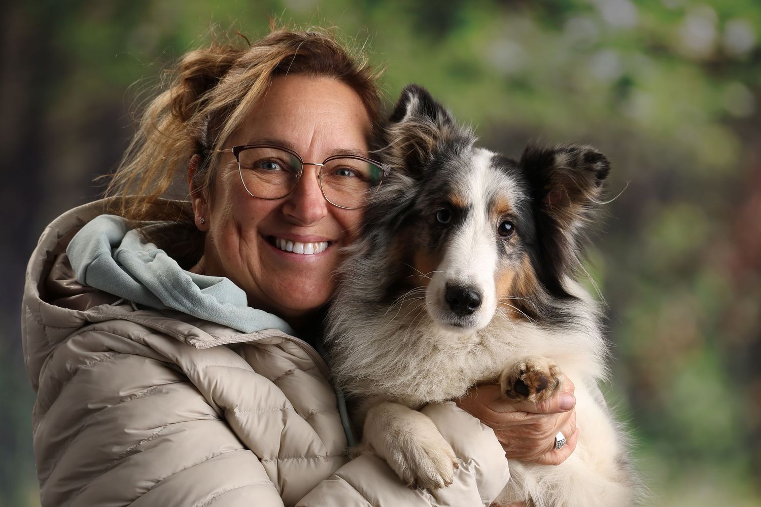 Smiling woman with glasses hugging a tri-color Australian Shepherd dog outdoors in a green forest setting.