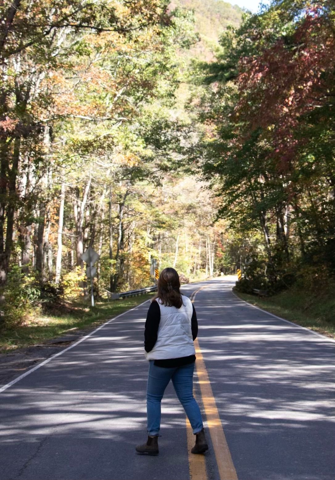 Woman standing on scenic autumn tree-lined road surrounded by colorful fall foliage.