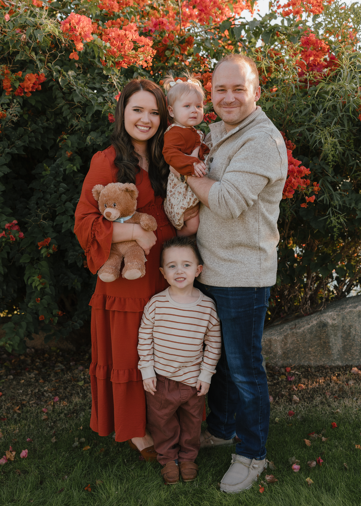 A young family of four poses happily in front of vibrant red bougainvillea blooms.