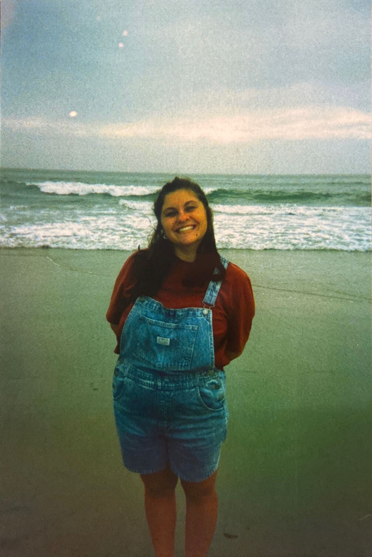 Smiling woman in denim overalls and red shirt stands on sandy beach with ocean waves behind her.