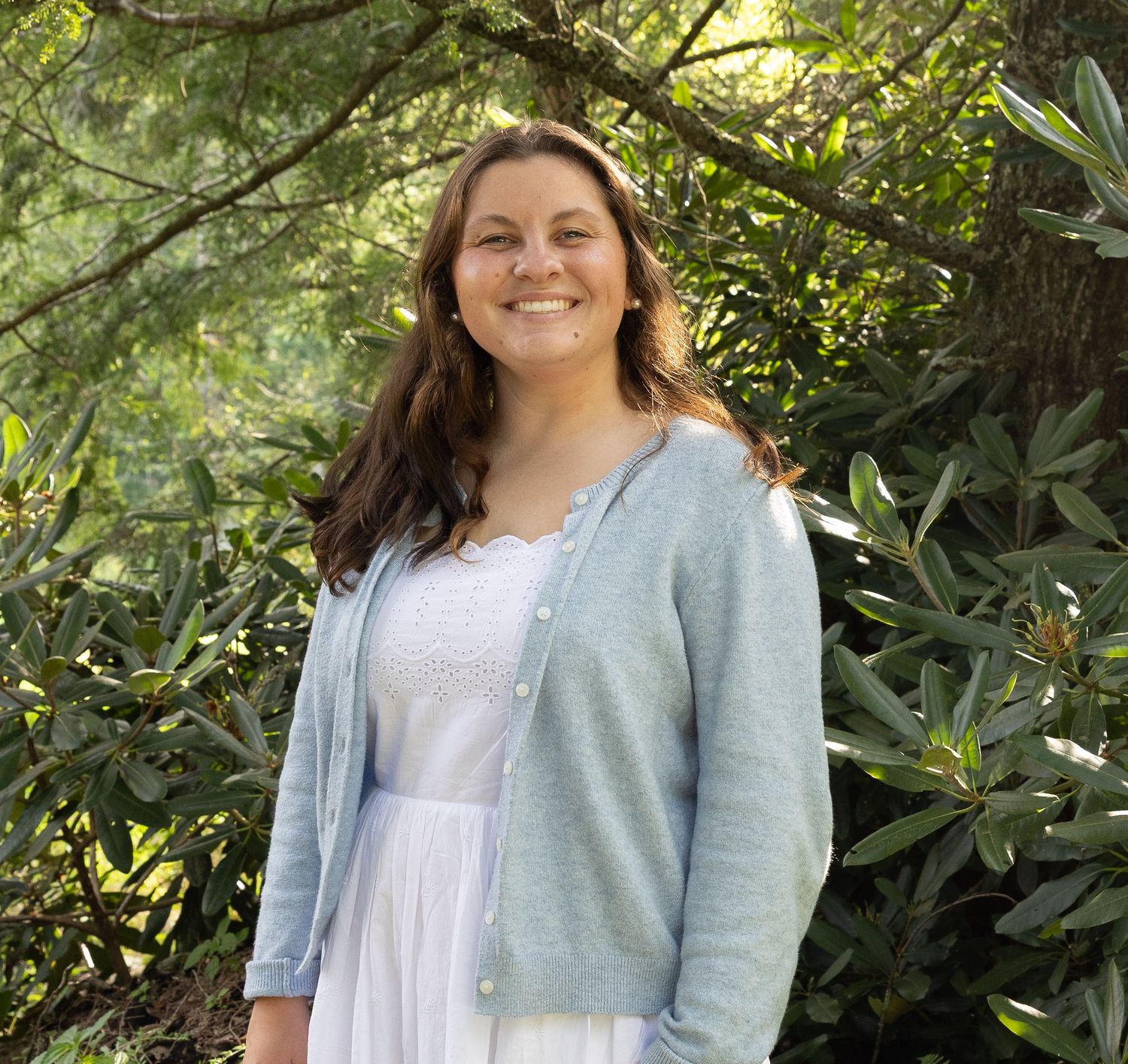 Smiling woman in white dress and blue cardigan standing outdoors among lush green trees.