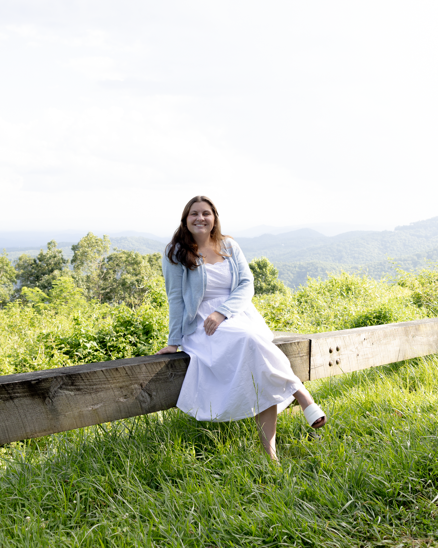 Woman in white dress and blue cardigan sitting on a wooden fence with green hills behind her.