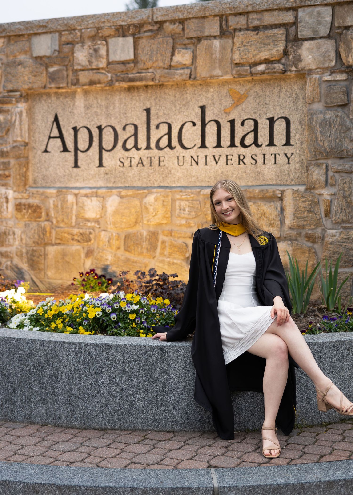 A smiling graduate in black robe and gold scarf sits by Appalachian State University stone sign surrounded by colorful flowers.