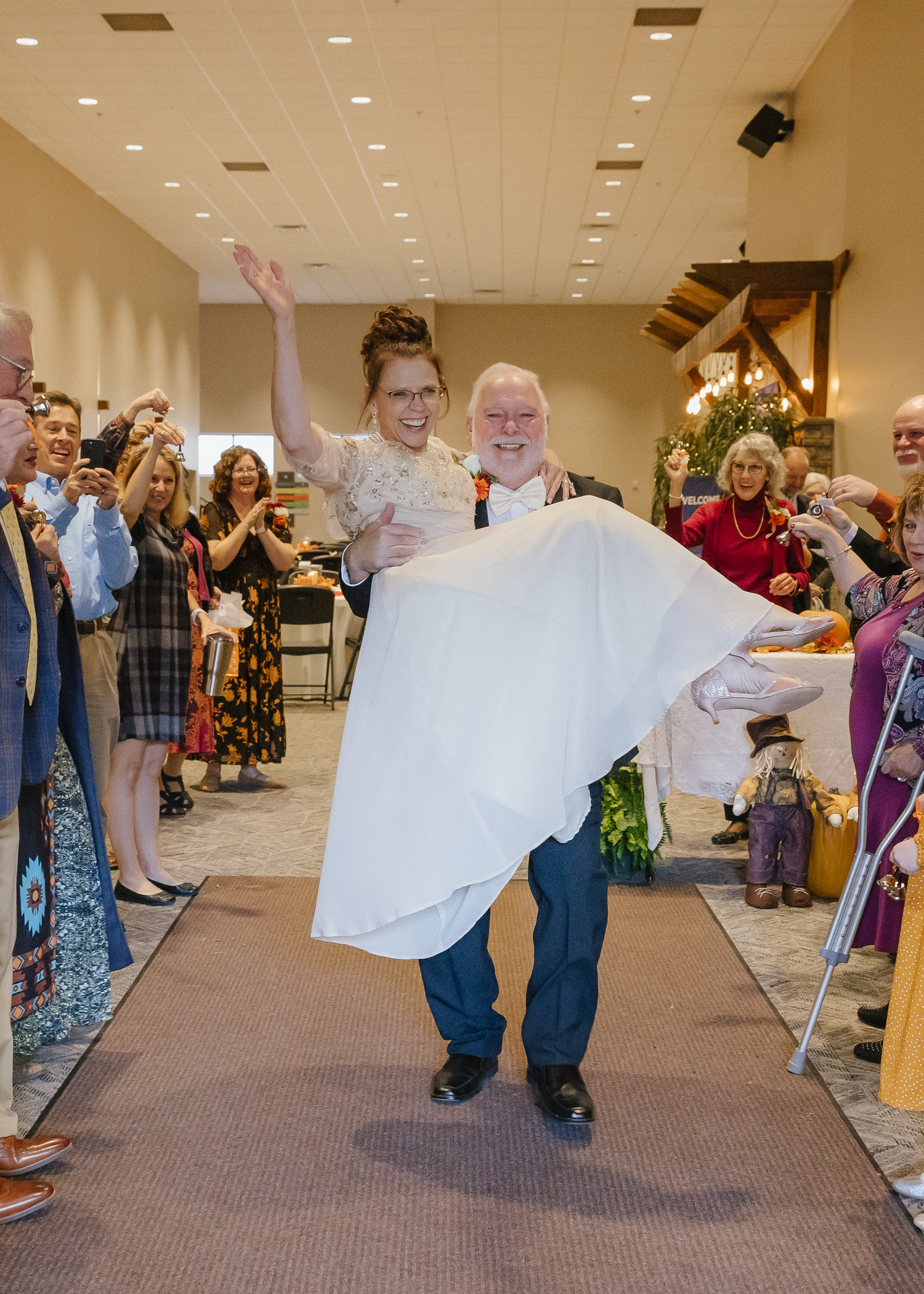 Groom carries joyful bride in white dress down reception aisle surrounded by cheering wedding guests.