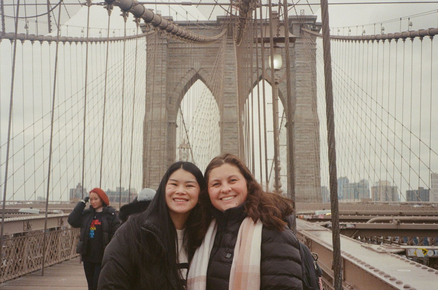 Two smiling women pose together on the Brooklyn Bridge on a cloudy day.