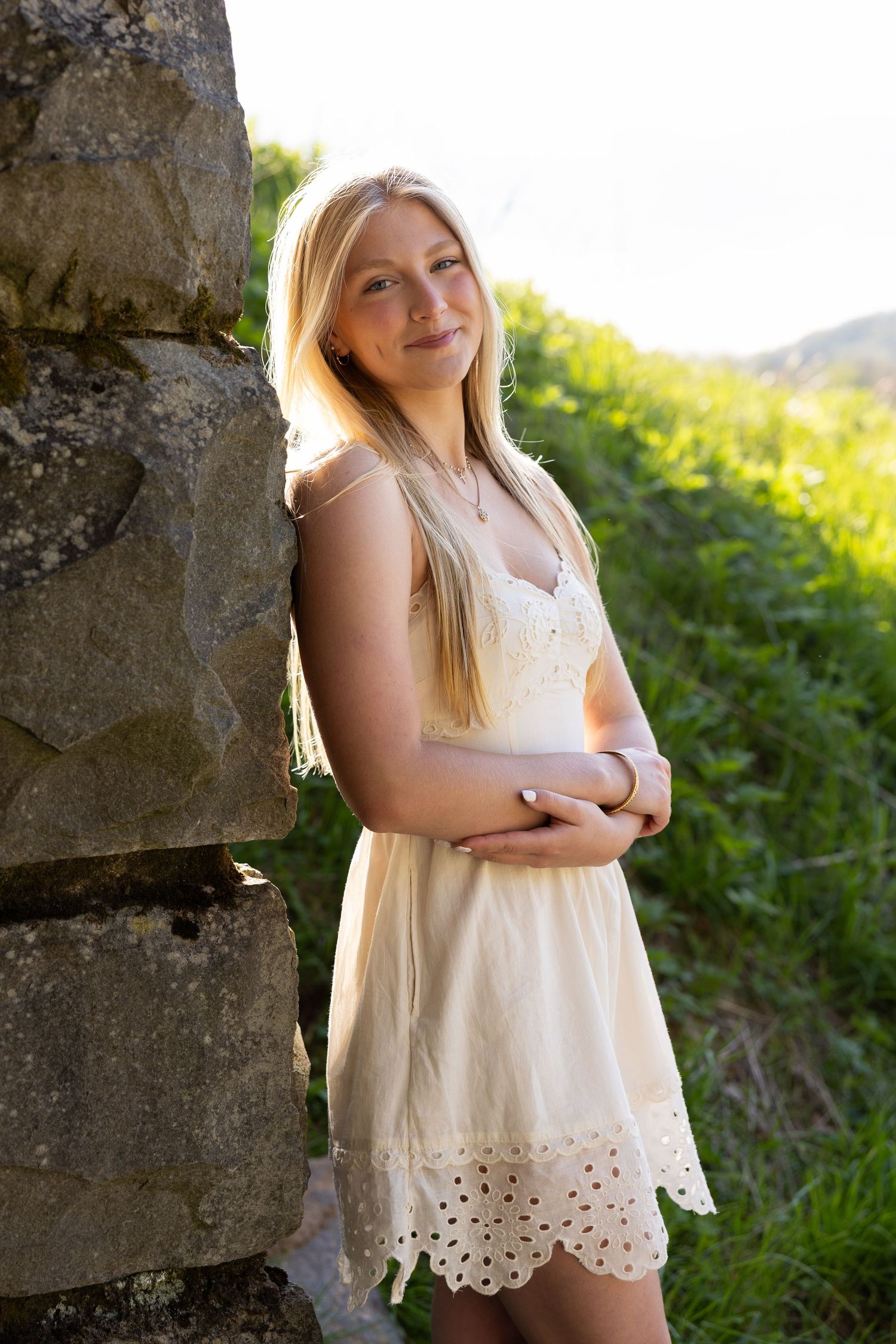 Young blonde woman in white sundress leaning against stone wall surrounded by green grass in golden sunlight.