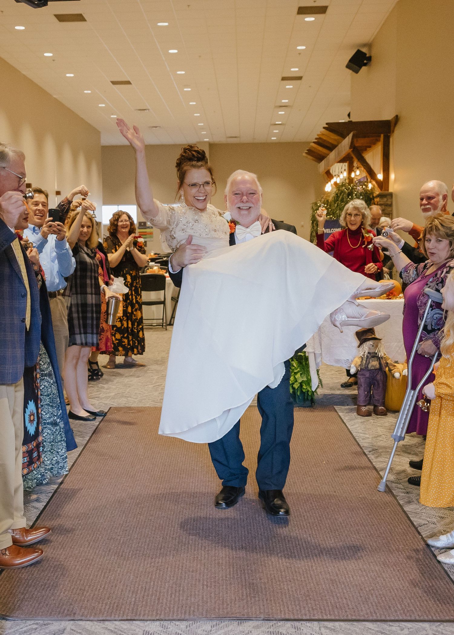 A groom carries his bride down the aisle as wedding guests celebrate around them indoors.