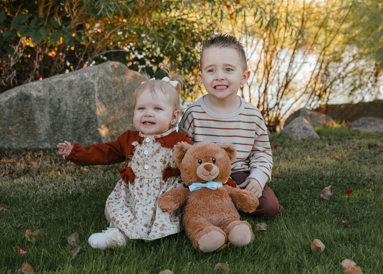 Two young siblings sit on grass outdoors, holding a teddy bear with a blue bow tie.
