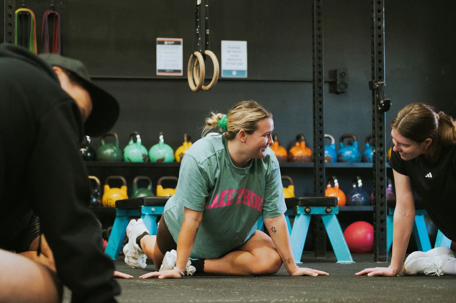 Woman in green Dayton shirt stretching on gym floor near kettlebell rack with instructor.