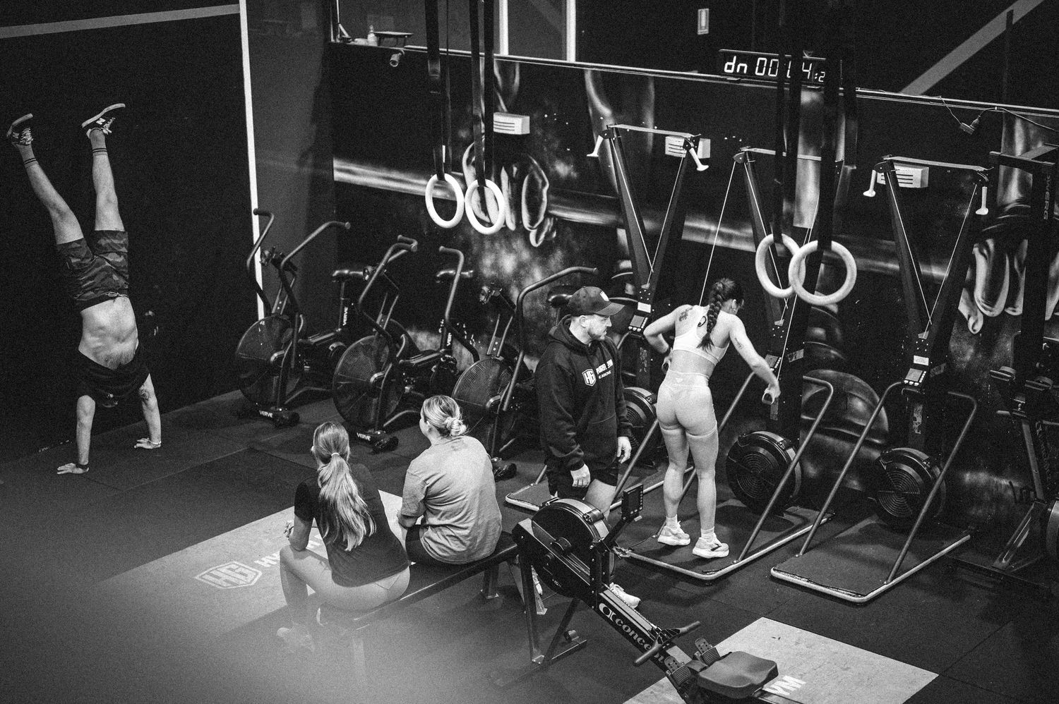 Black and white photo of gym members training with rowing machines and gymnastics rings.