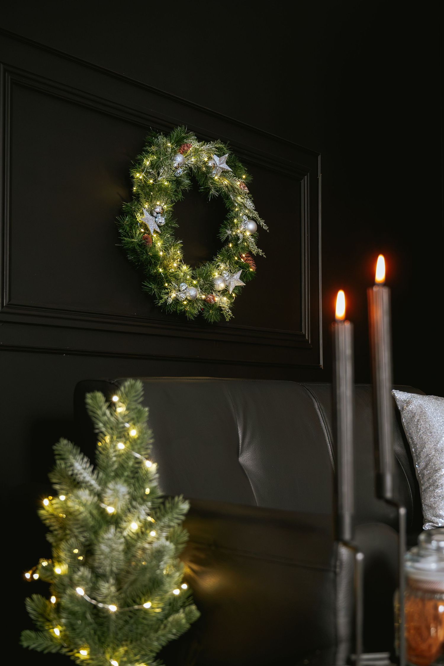 Lit Christmas wreath with silver stars hangs on a dark wall above a small lit tree, with tall black candles glowing beside a leather sofa.