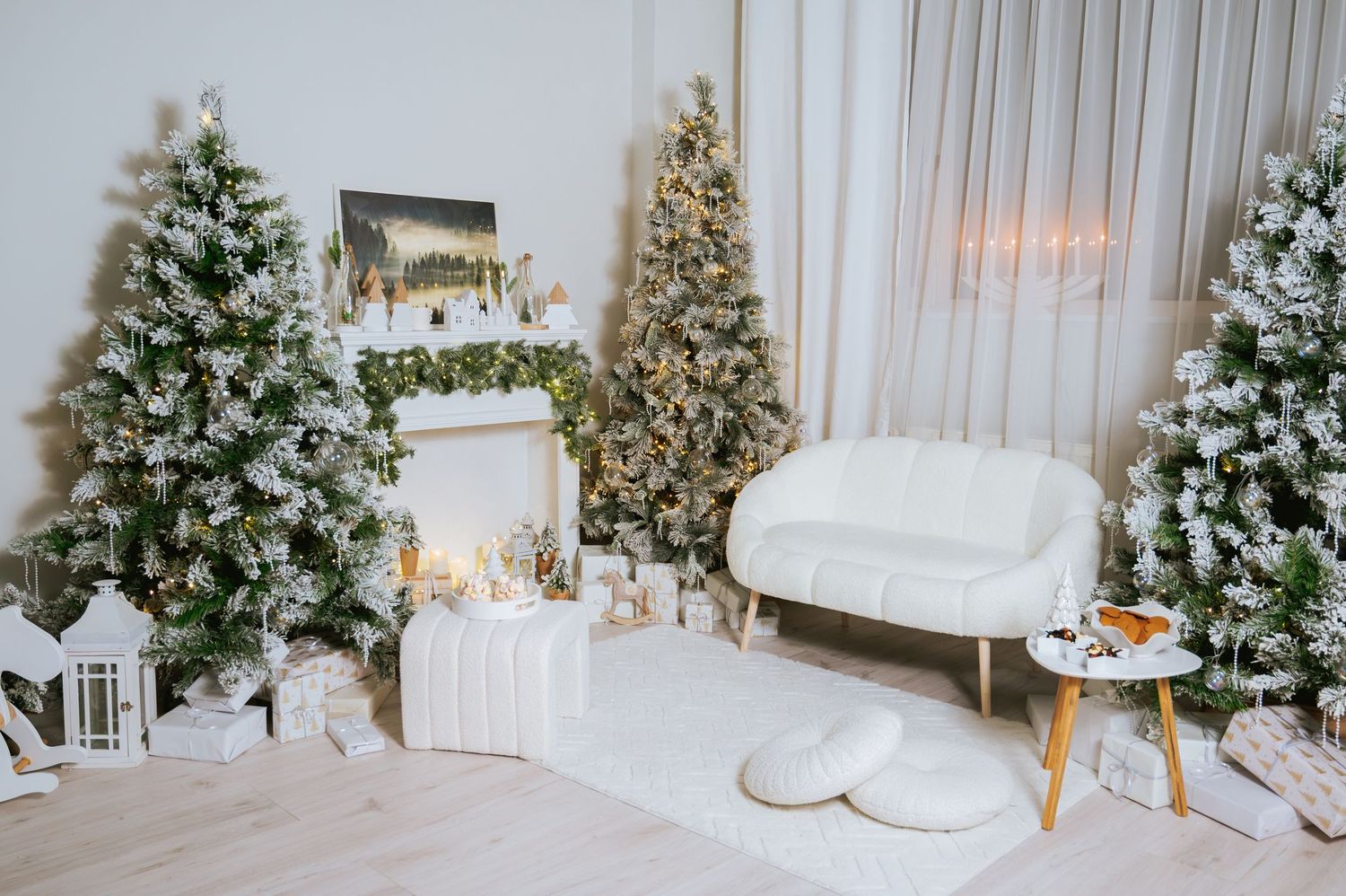 Cozy white Christmas living room with frosted trees, a decorated fireplace mantel, cream sofa, floor cushions, lanterns and wrapped gifts.