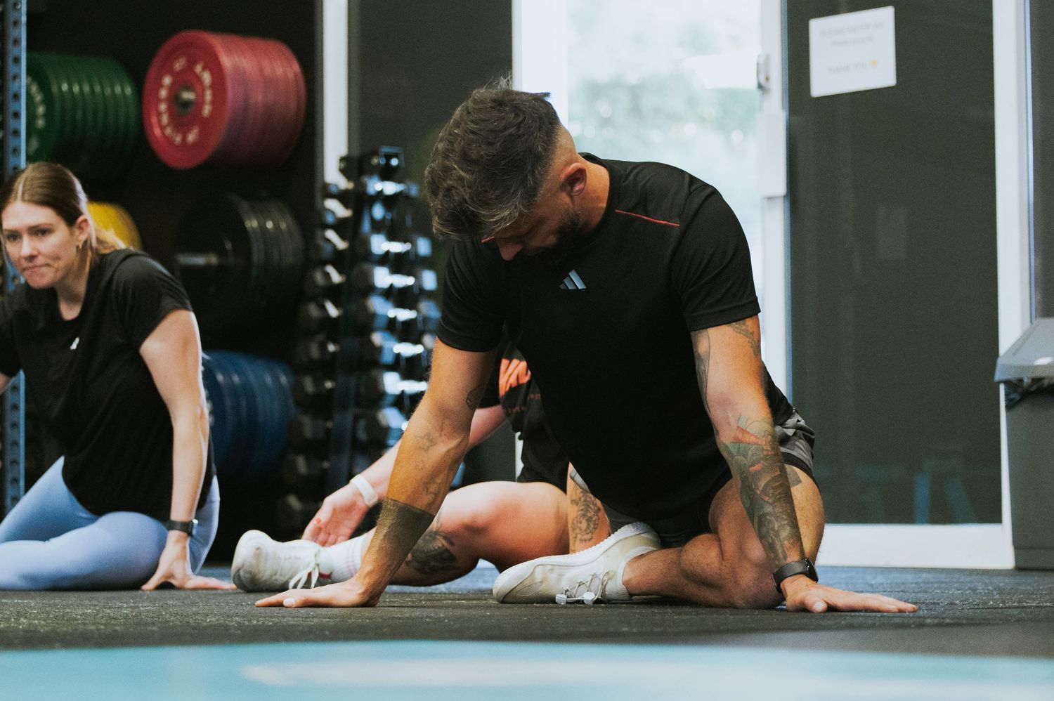 Fitness coach assisting a seated athlete with stretching on a gym floor mat.