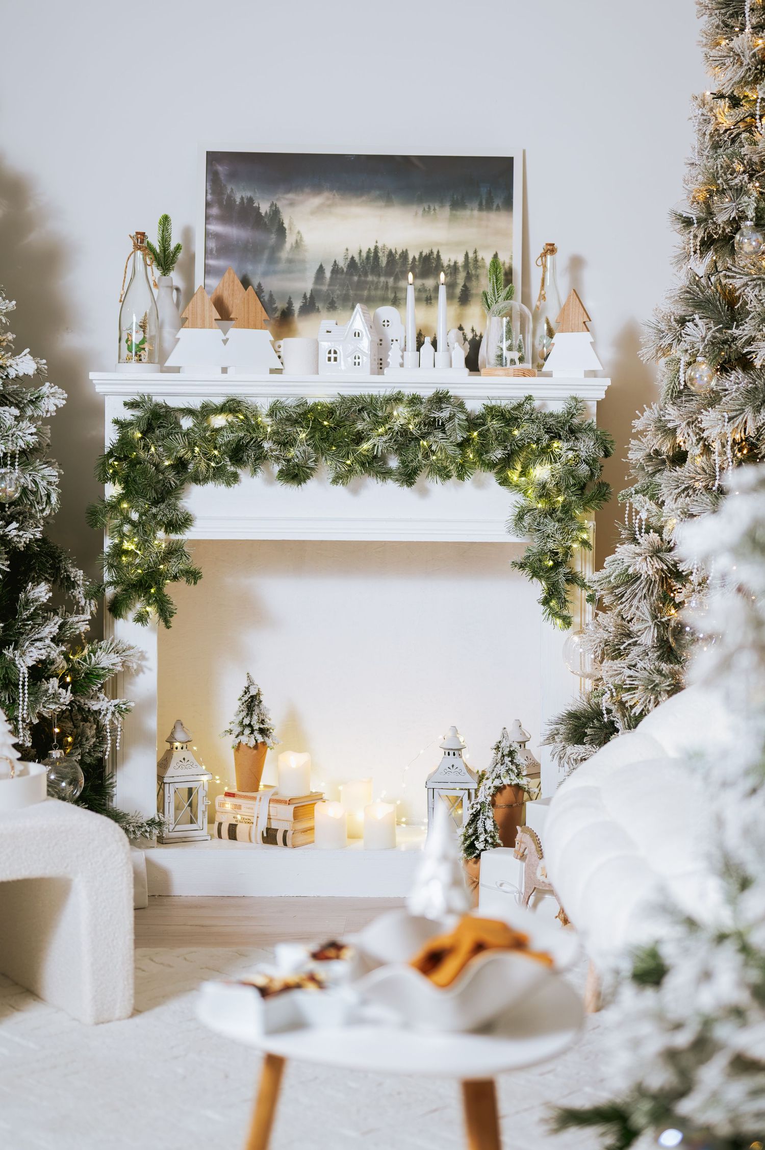 Cozy white Christmas living room with a faux fireplace mantel, greenery garland, flocked trees, candle lanterns and a wooden village décor.
