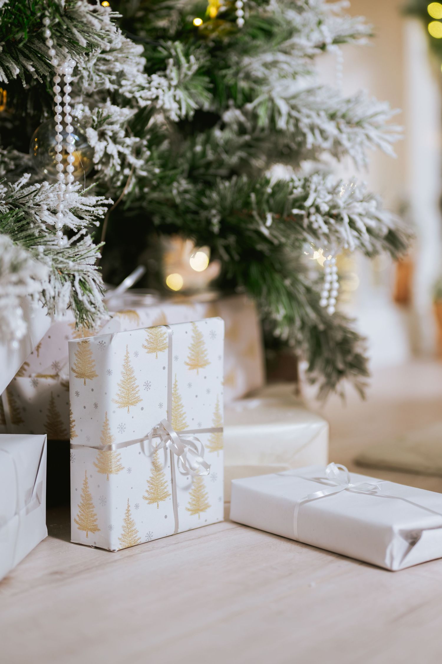 Gift boxes wrapped in white and gold Christmas tree paper with ribbon sit on a wood floor beneath a frosted, softly lit Christmas tree.