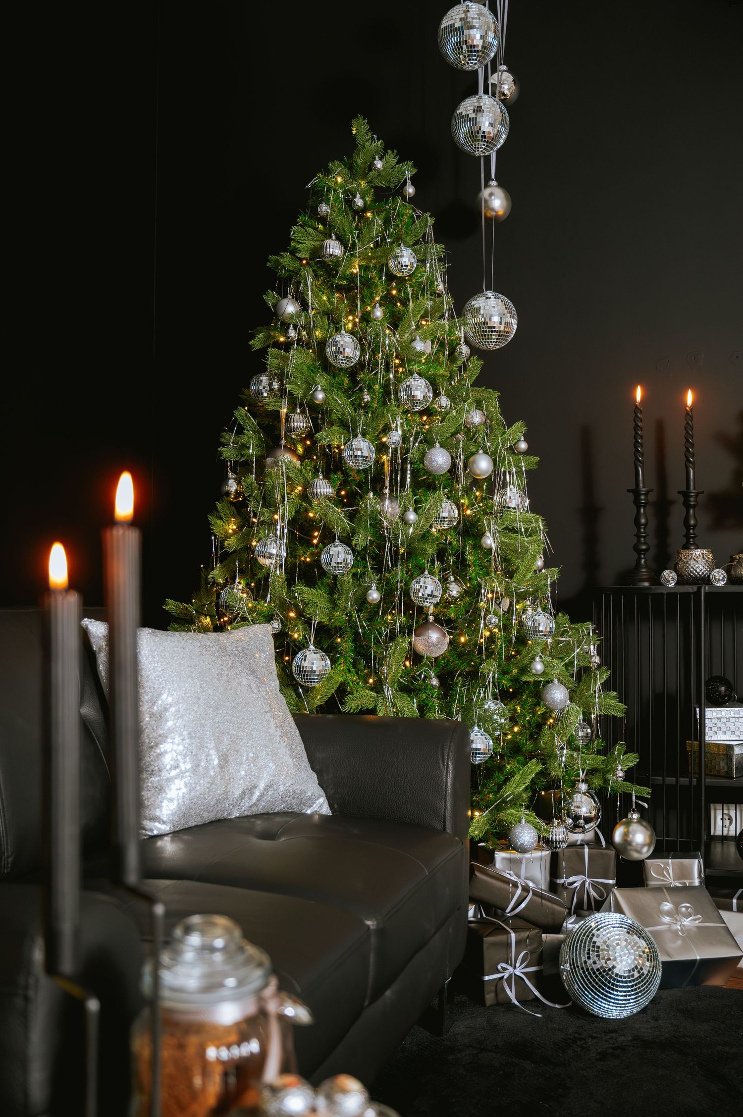 Christmas tree with silver ornaments and string lights beside a black leather sofa, lit candles, and wrapped gifts in a dark living room.