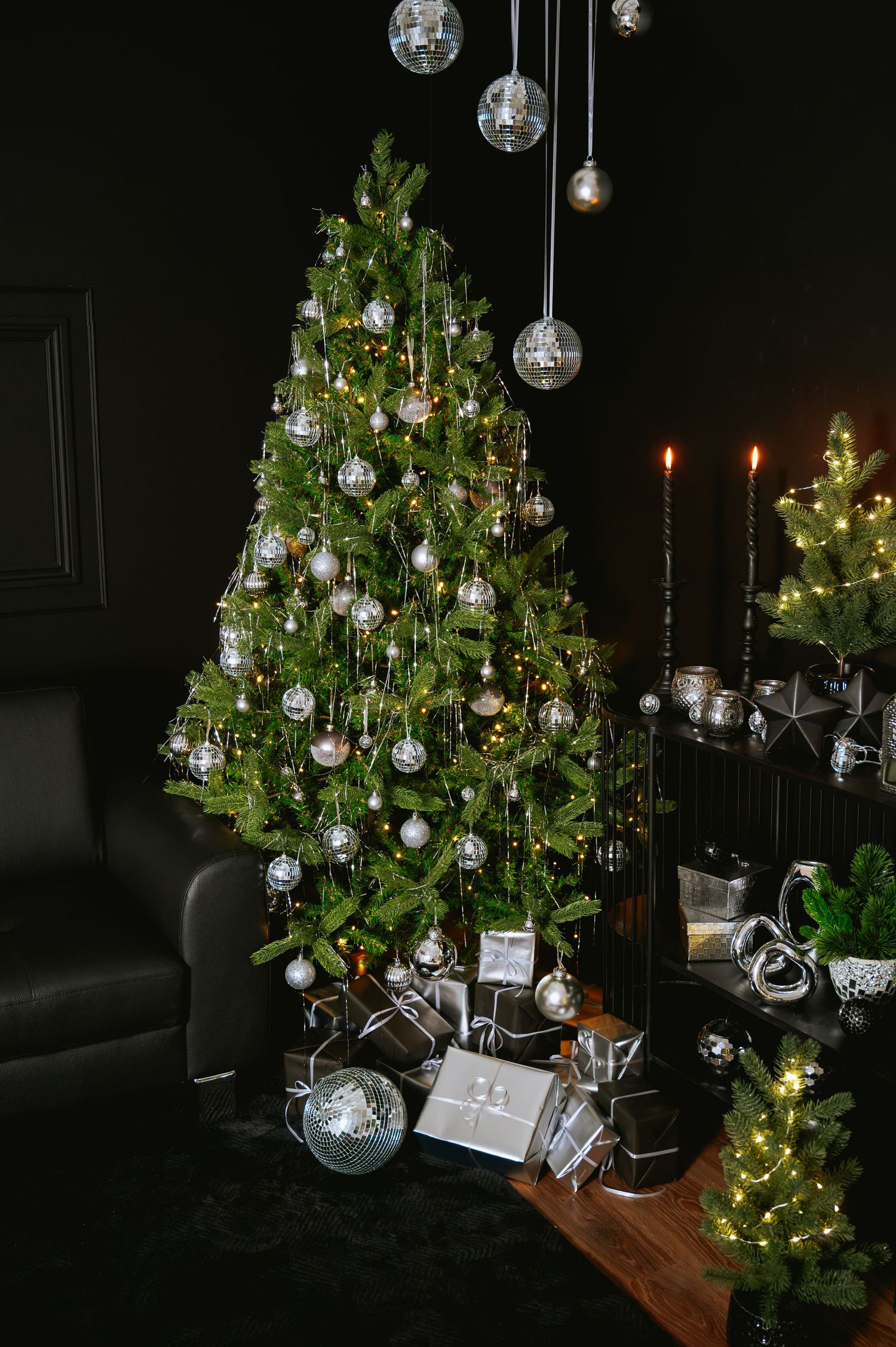 Silver and white decorated Christmas tree with mirrored disco balls, wrapped gifts, black candles and small lit trees in a dark, moody room.