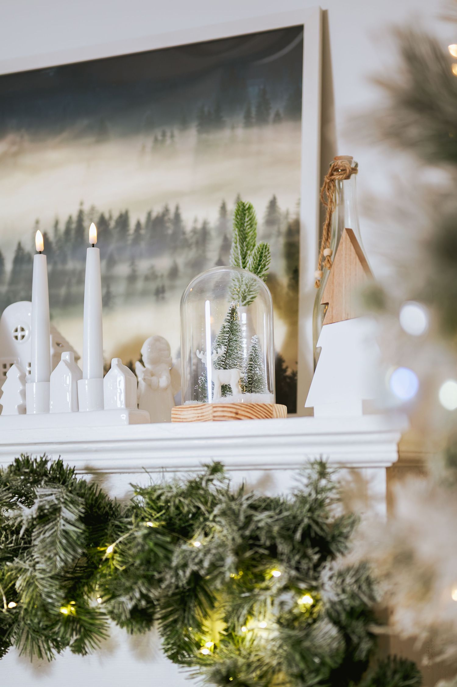 Christmas mantel decor with lit white candles, mini ceramic houses, a glass cloche with a tiny tree, and a lighted evergreen garland.