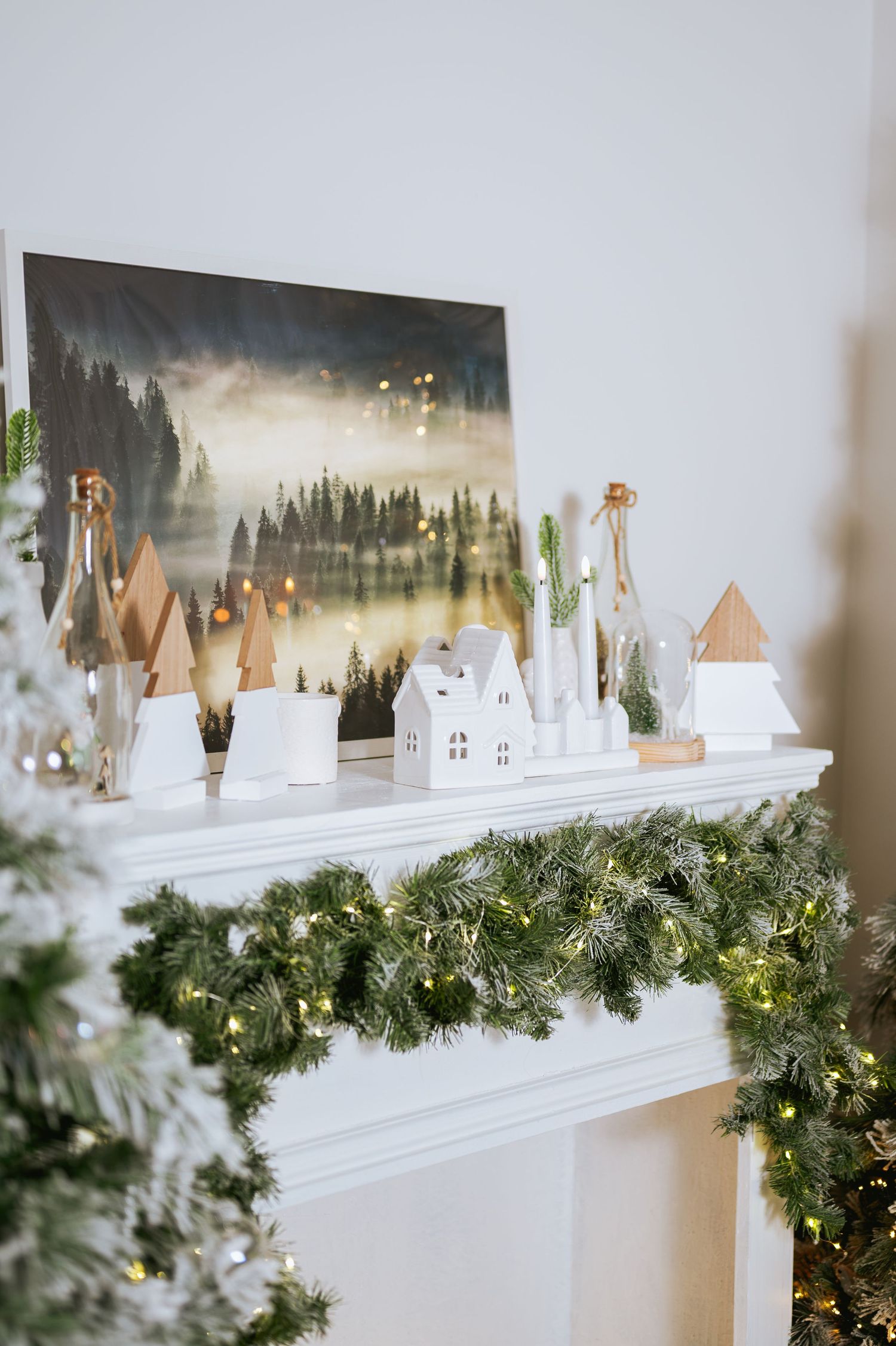 White fireplace mantel decorated with a frosted evergreen garland, string lights, miniature white houses and a forest landscape painting.