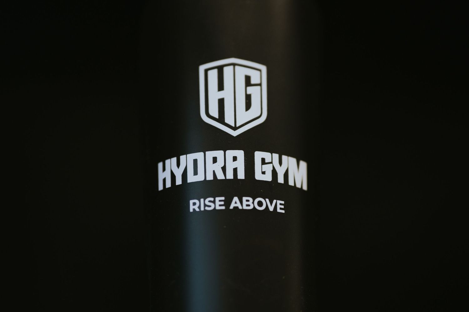 Black metal water bottle featuring HG Hydra Gym Rise Above logo in silver on dark background.