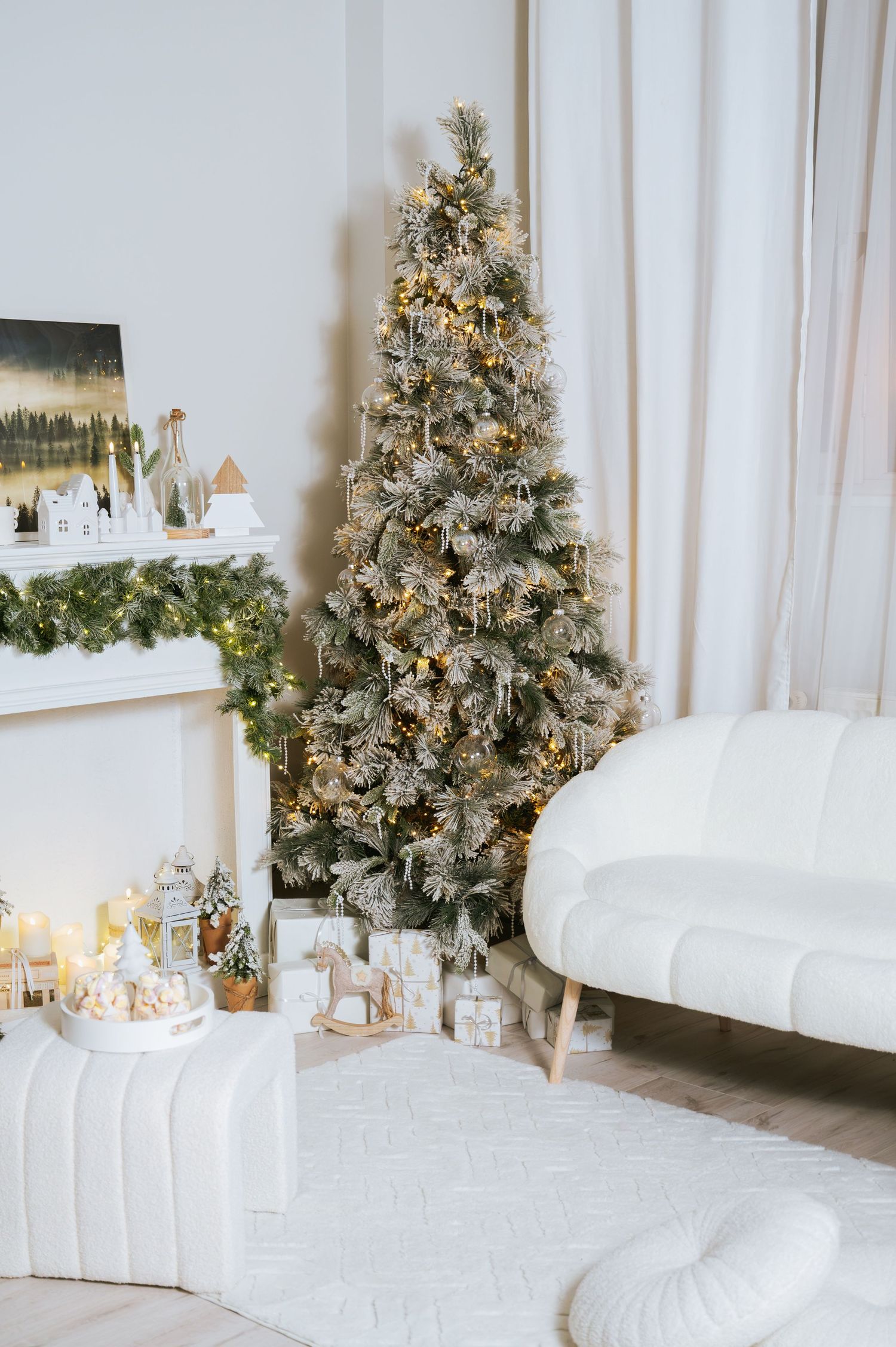Cozy white living room with a flocked Christmas tree, warm string lights, a garland-draped mantel, wrapped gifts and a curved boucle sofa.