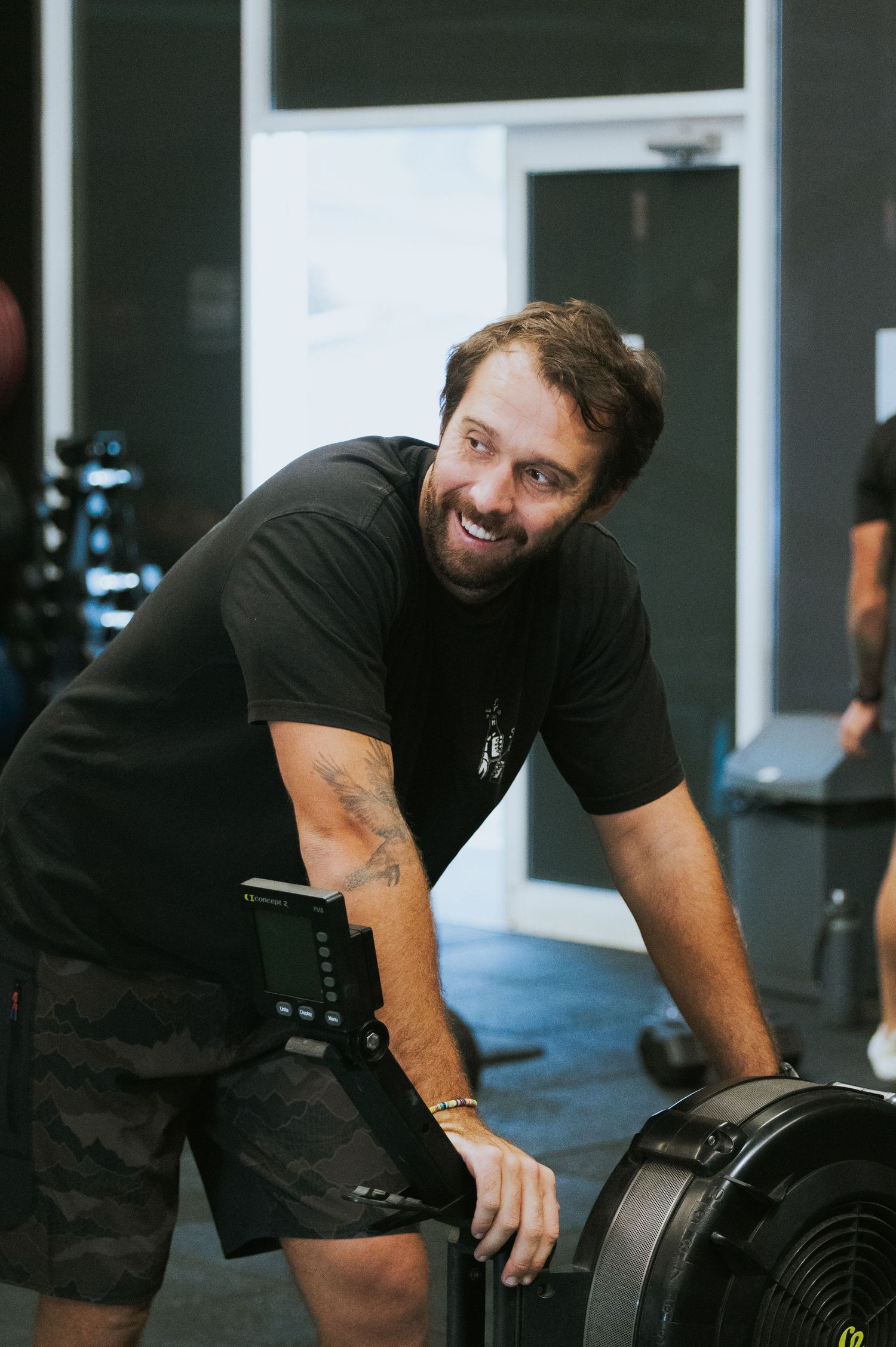 Smiling bearded man in black shirt leaning on a rowing machine at a CrossFit gym.