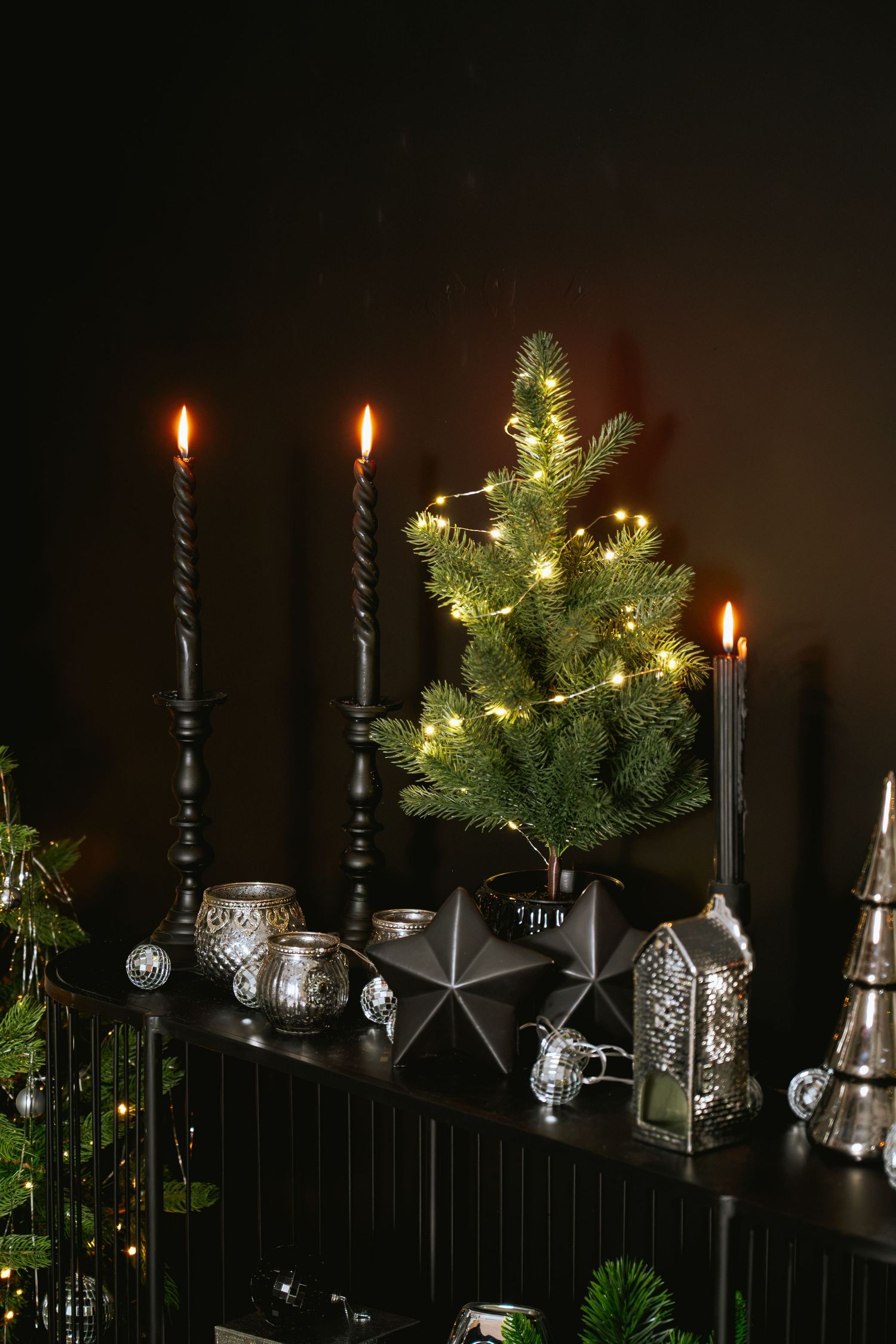 Small lit Christmas tree with string lights beside black twisted candles, metallic stars and silver ornaments on a dark shelf.