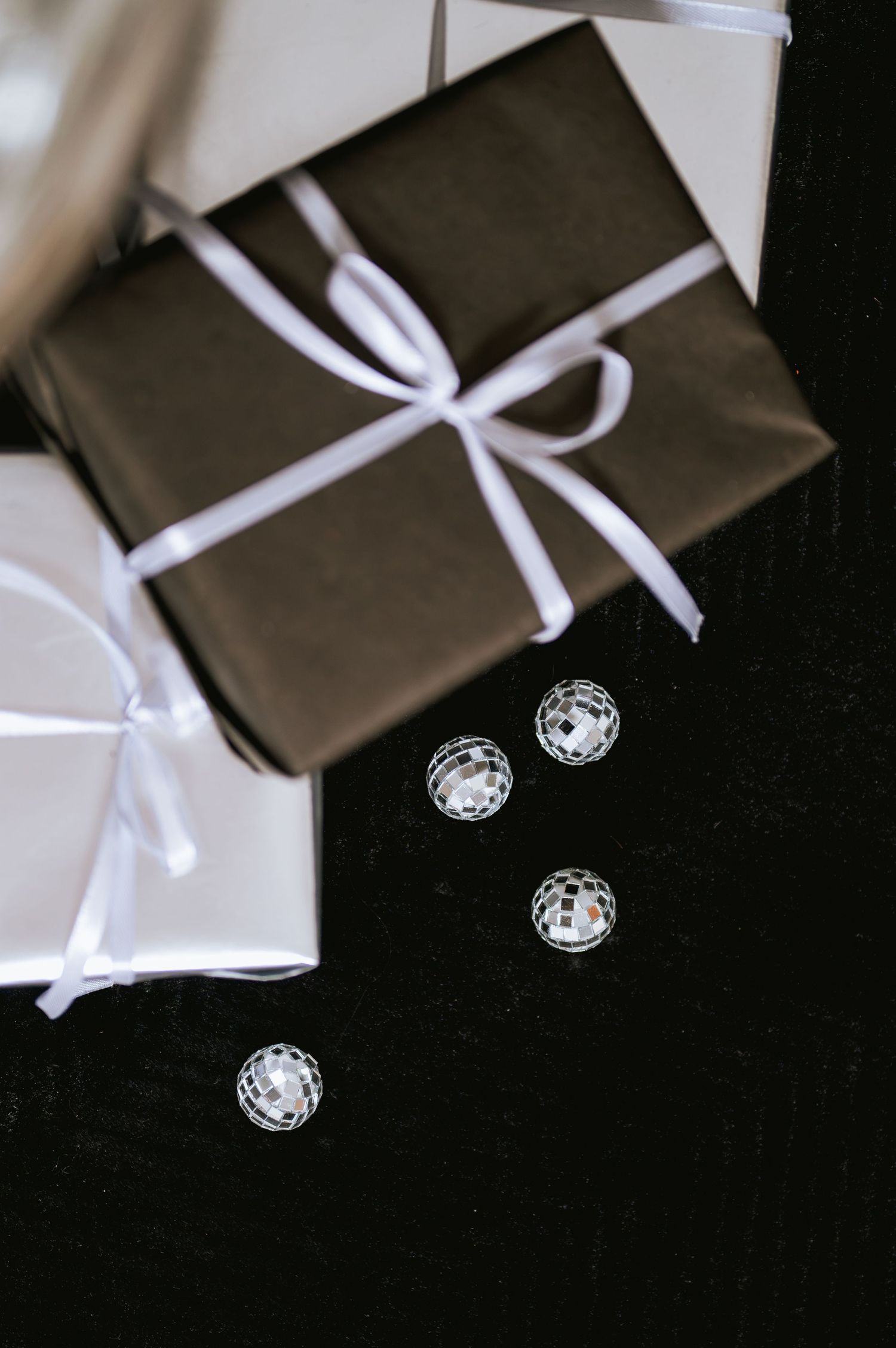 Overhead view of elegant gift boxes wrapped in black and silver paper with white satin ribbons beside small mirrored disco ball ornaments.