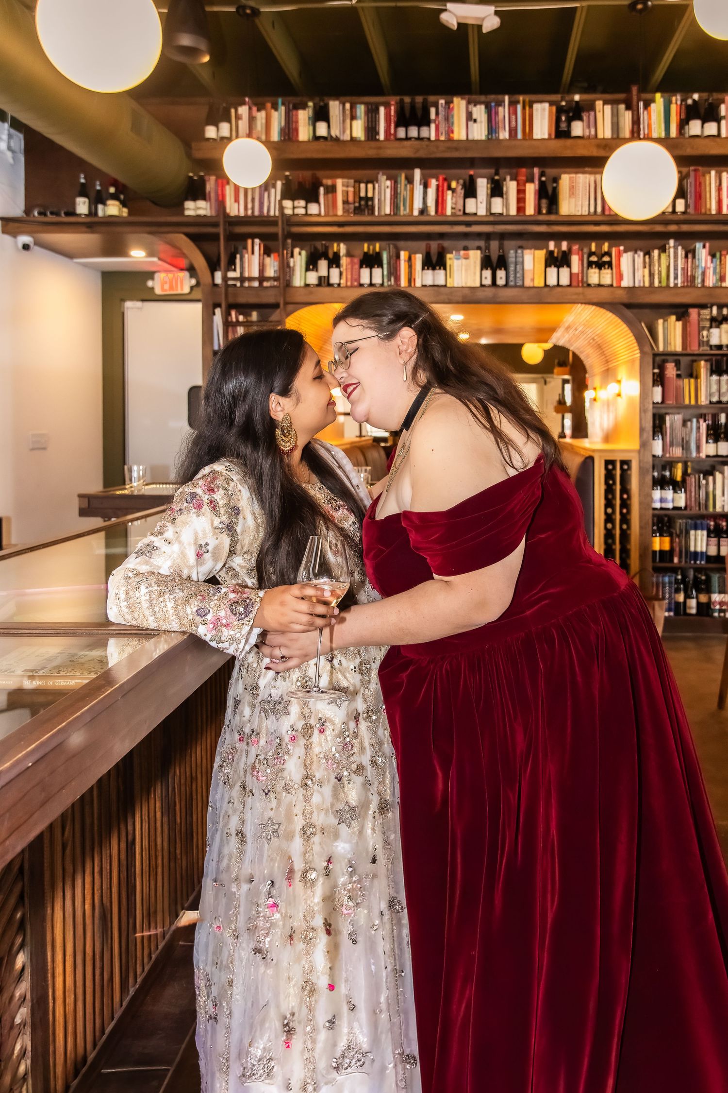 Two women sharing a kiss at a wine bar with bookshelves in background.