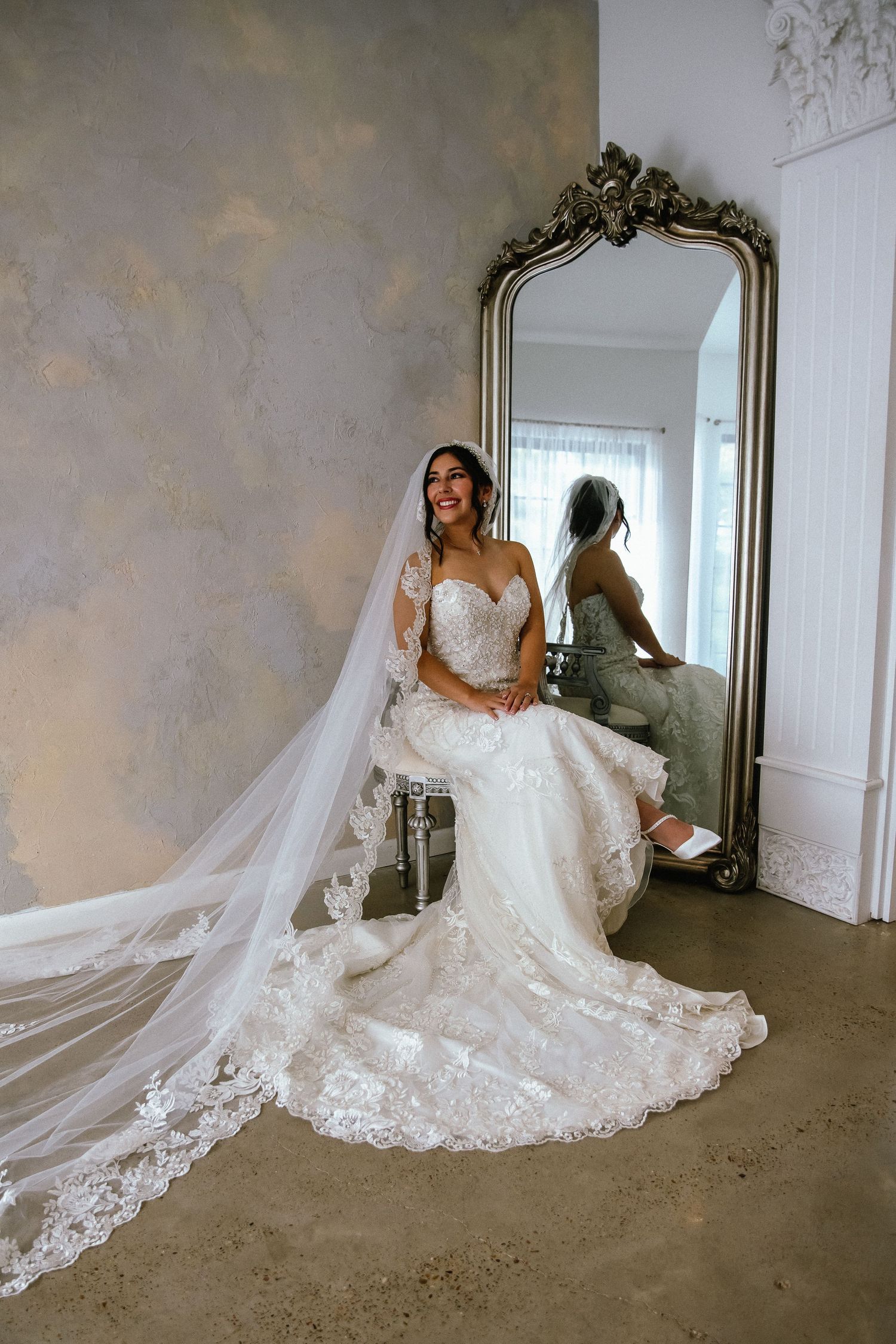 Smiling bride in lace wedding gown with cathedral veil seated by ornate mirror.