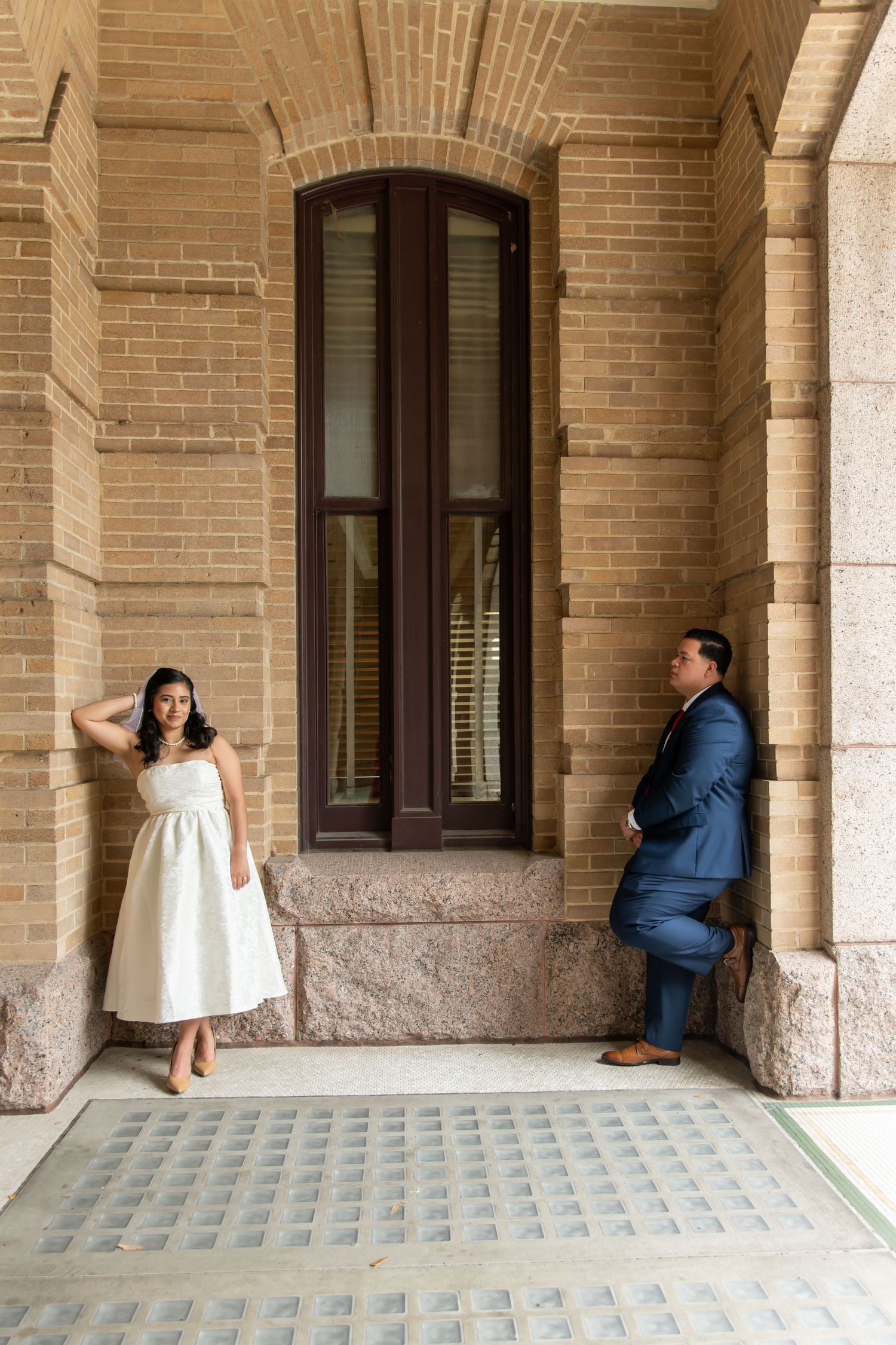 Bride in tea-length white dress and groom in blue suit pose separately by ornate brick building window.