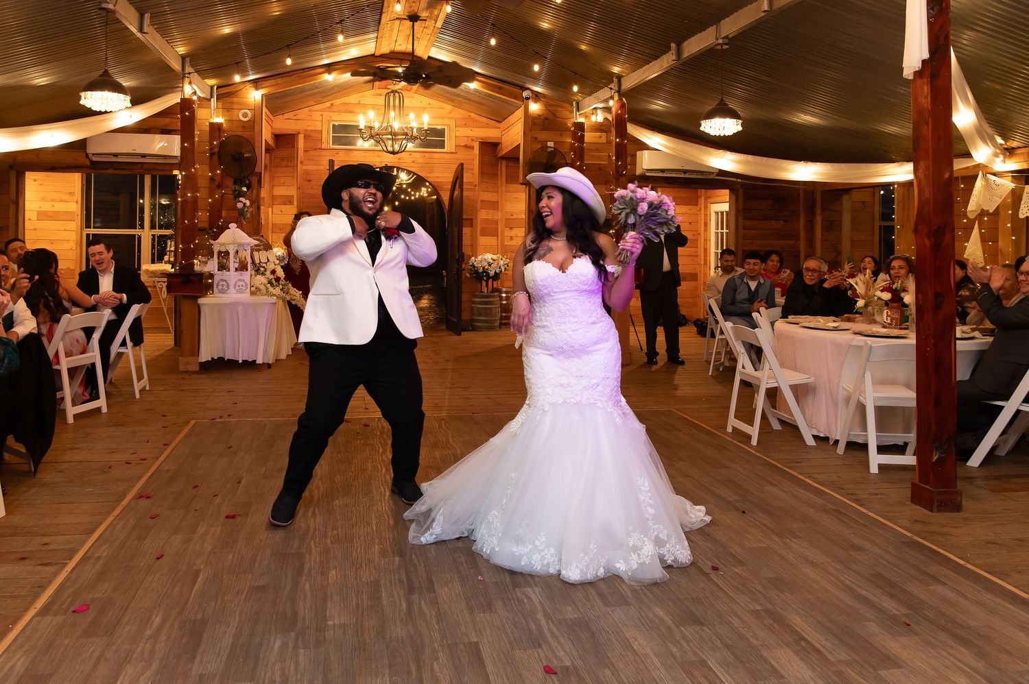 Bride and groom in cowboy hats share a lively first dance in a warmly lit rustic barn reception.