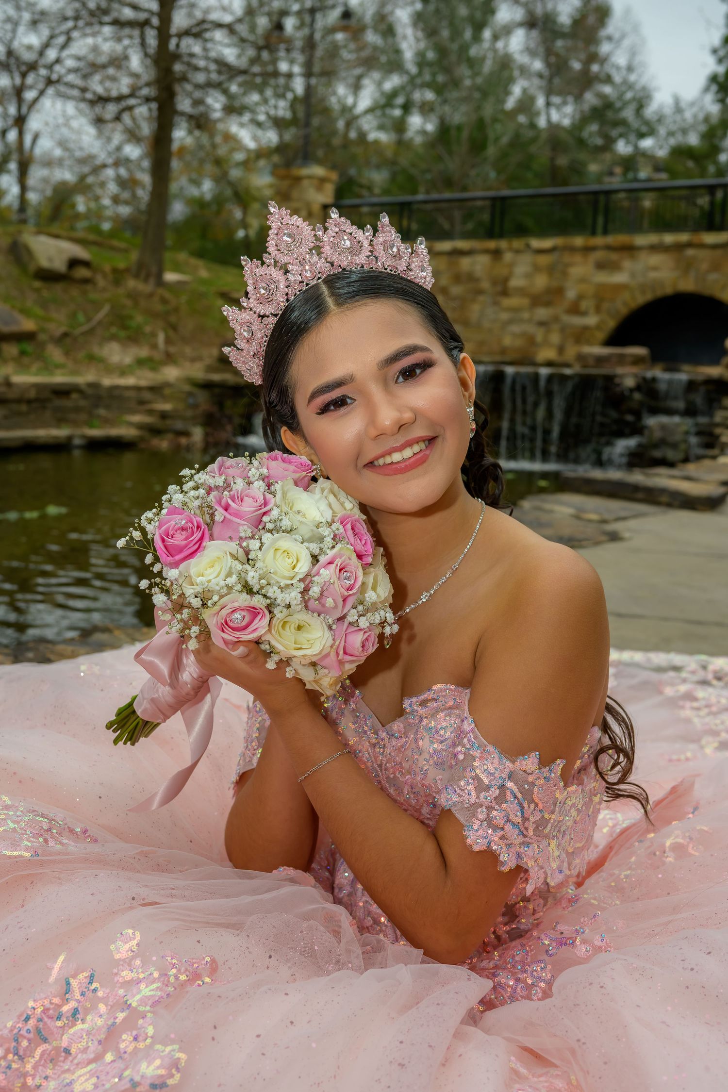Smiling young woman in pink quinceañera gown and crown holds a bouquet of pink and white roses outdoors.
