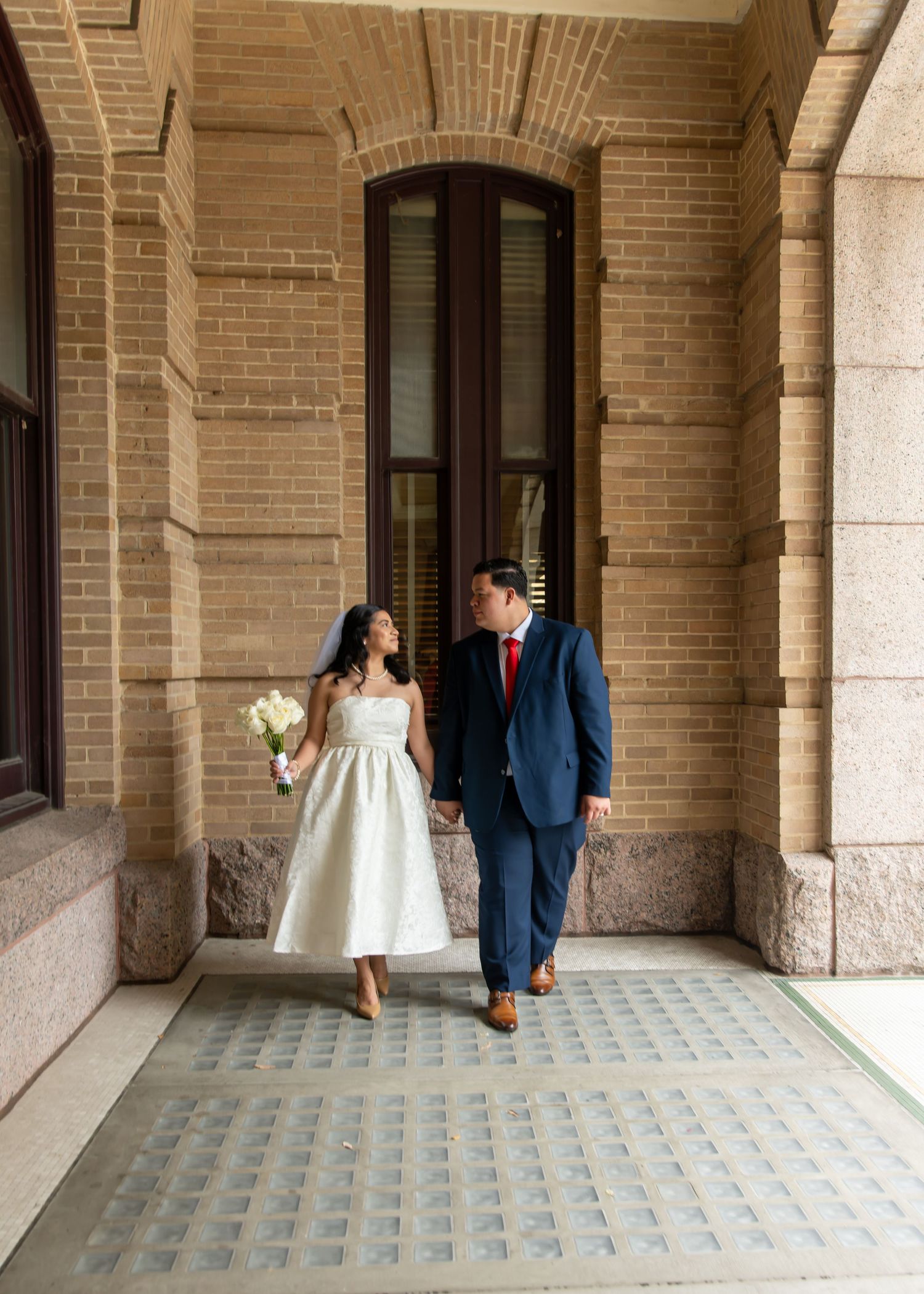 Bride holding white bouquet and groom in blue suit walk hand-in-hand along elegant brick building corridor.