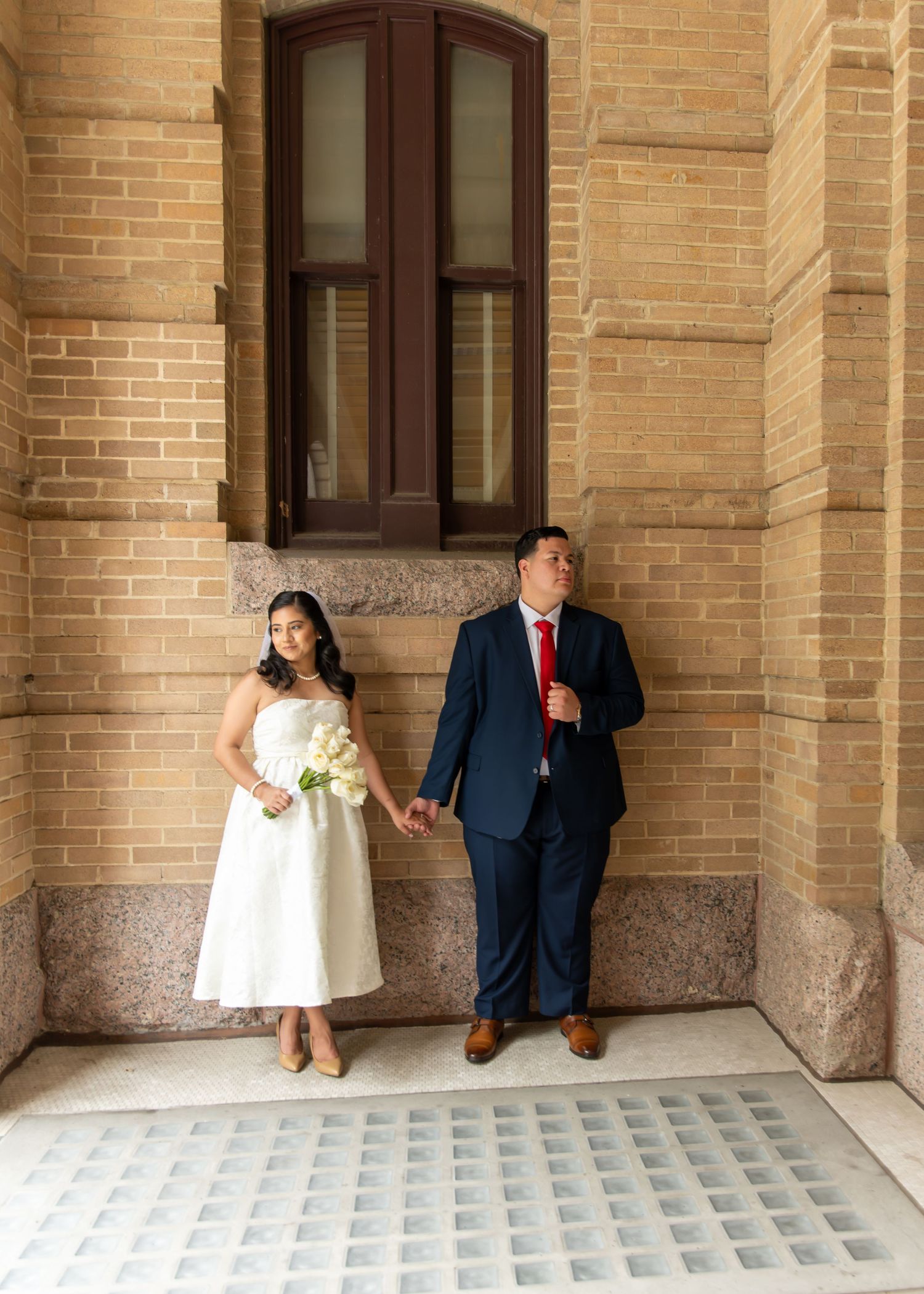 Bride with bouquet and groom in blue suit with red tie stand hand-in-hand against a brick building.