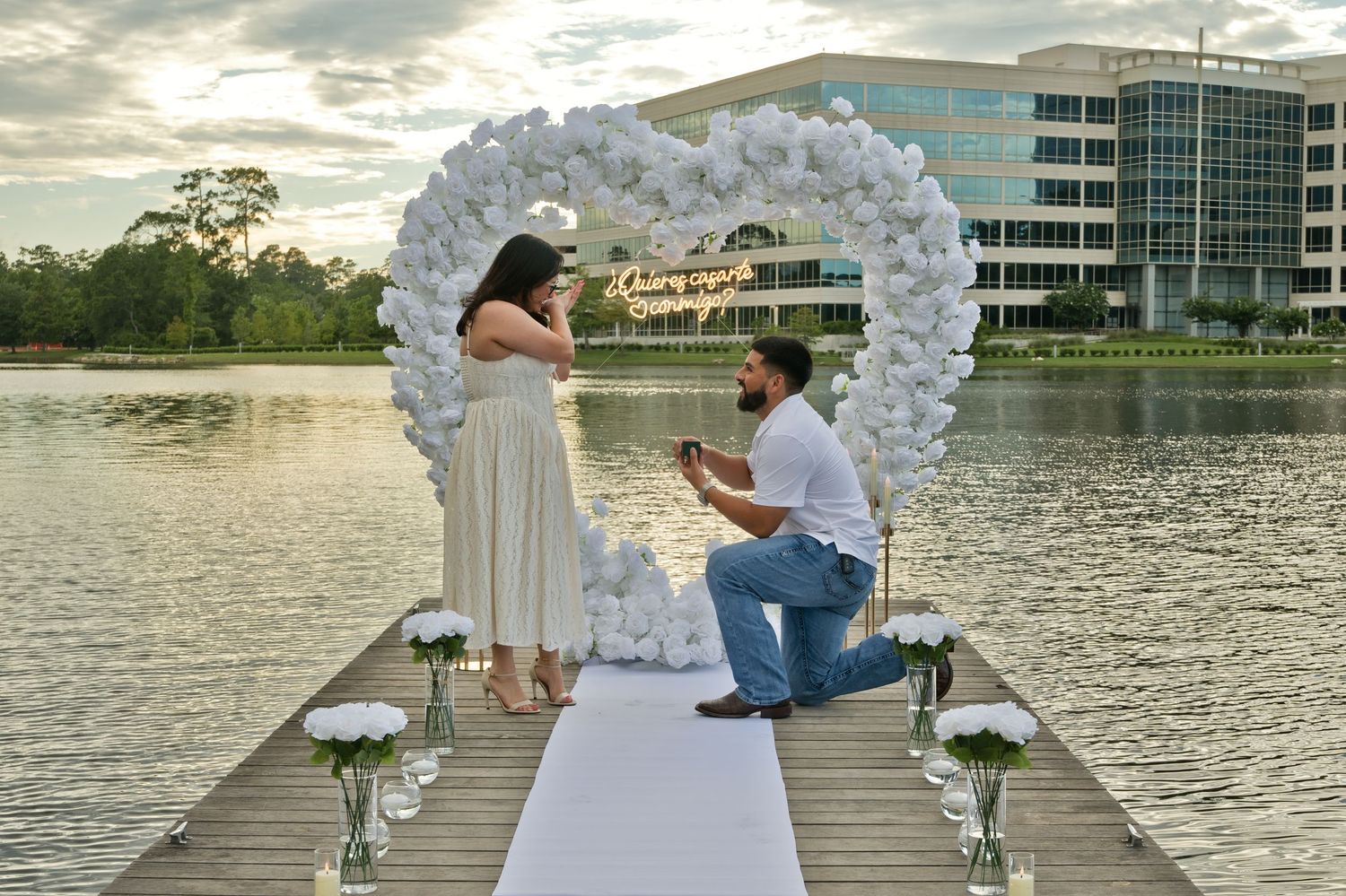 Man proposing on waterfront dock with white balloon heart arch and floral decorations at sunset.