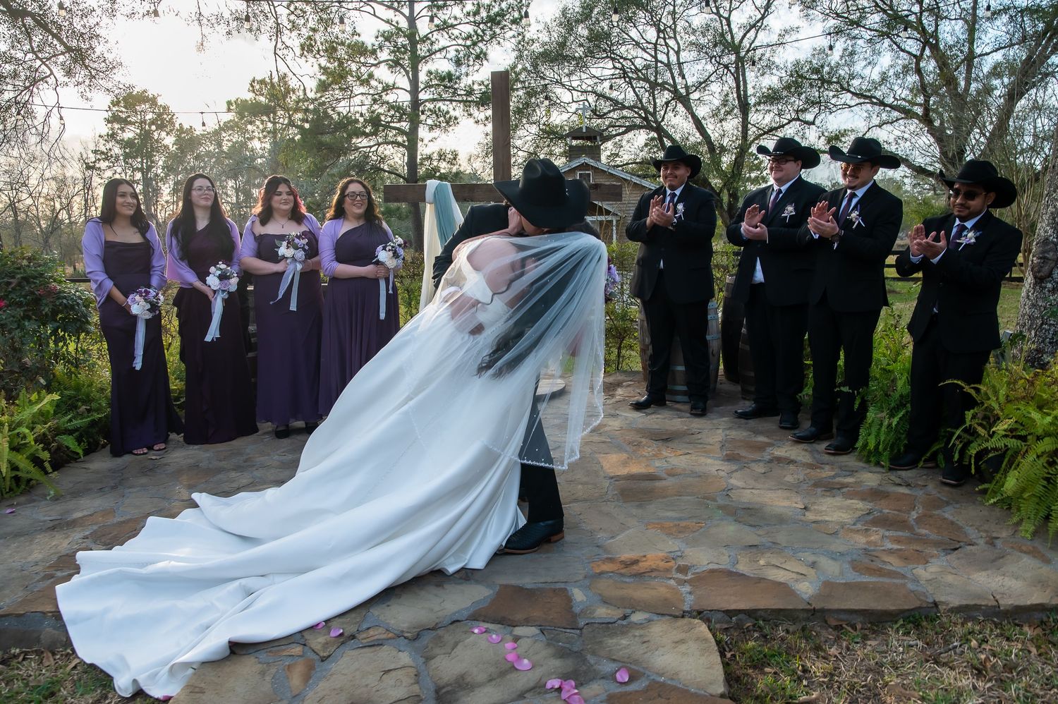 Bride and groom sharing a kiss while wedding party in purple dresses and cowboy hats applauds outdoors.