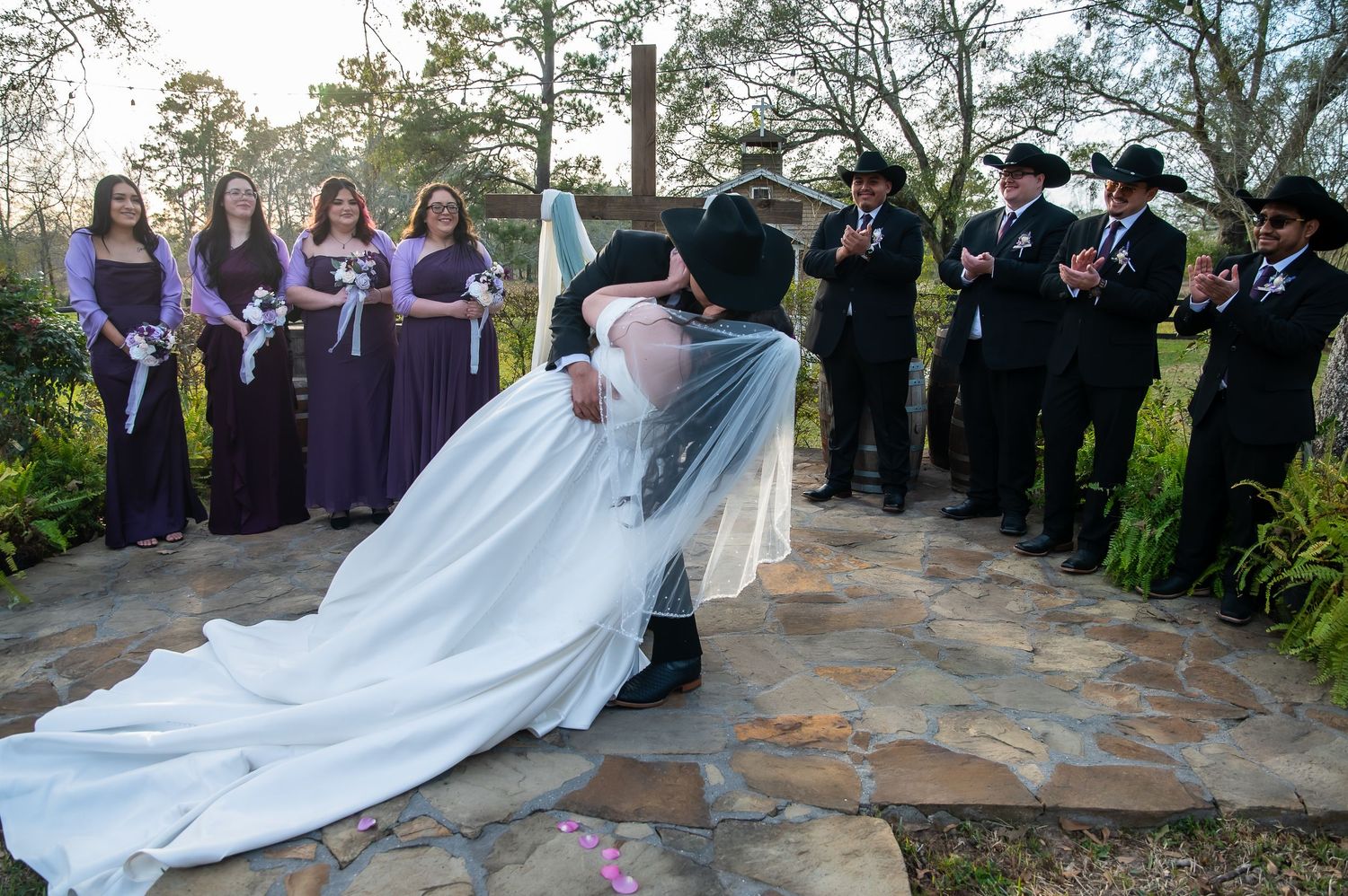Wedding party celebrates as groom dips bride in romantic kiss, bridesmaids in purple and groomsmen in black surround them.