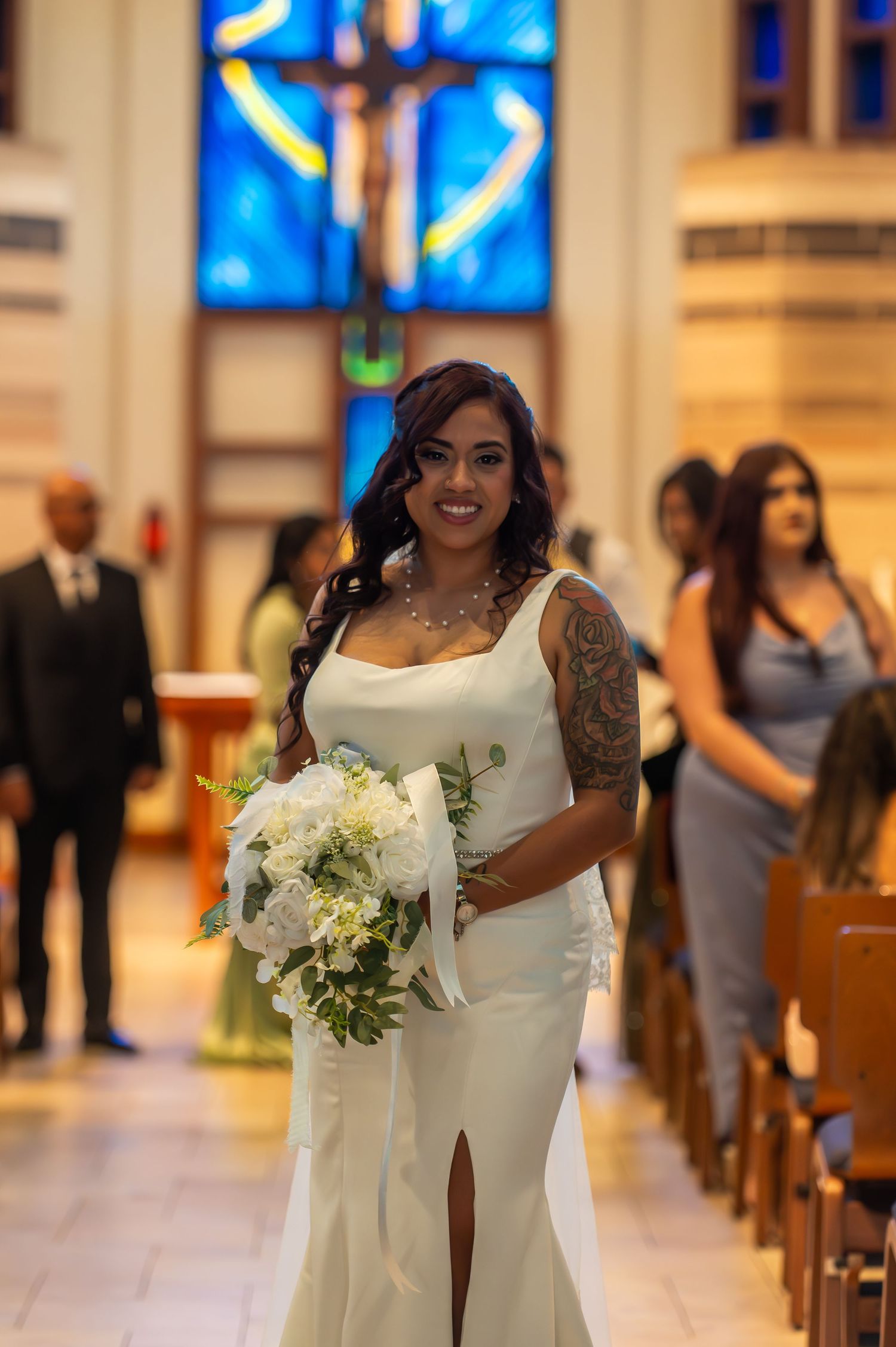 Bride in white fitted gown with floral tattoo carrying bouquet walking down church aisle.