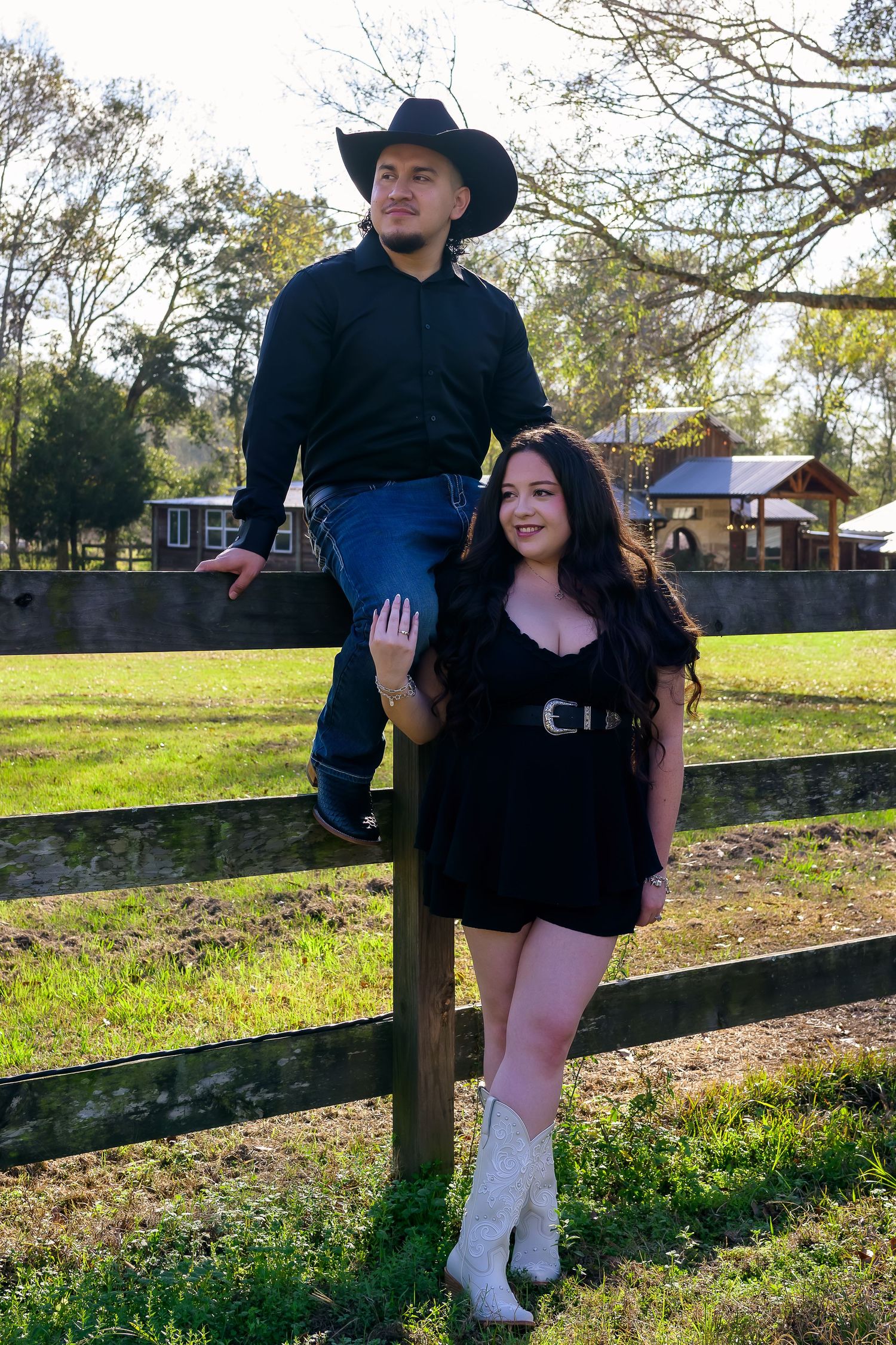 Couple in western attire posing at wooden fence on a sunny farm.