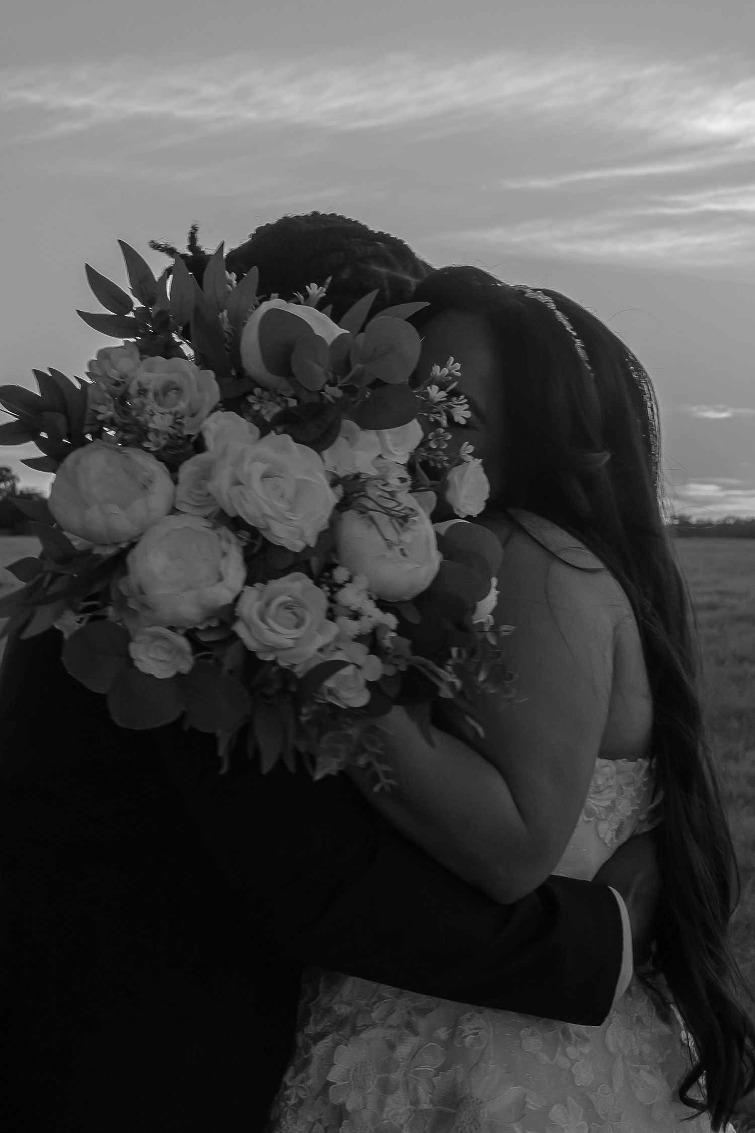 Black and white photo of couple kissing behind bridal bouquet at sunset.